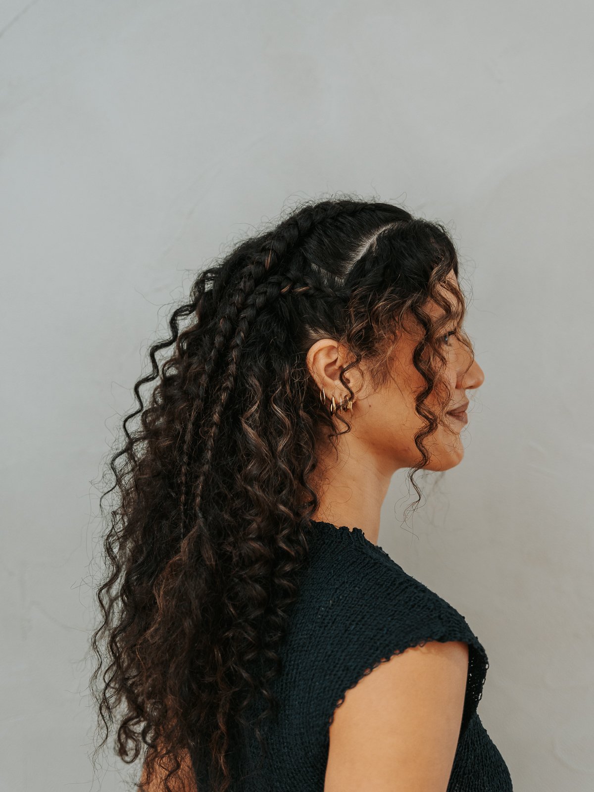 Side profile of a woman with curly brown hair styled with braids on top and loose curls down her back, wearing gold hoop earrings and a black textured top, standing against a plain light gray background.