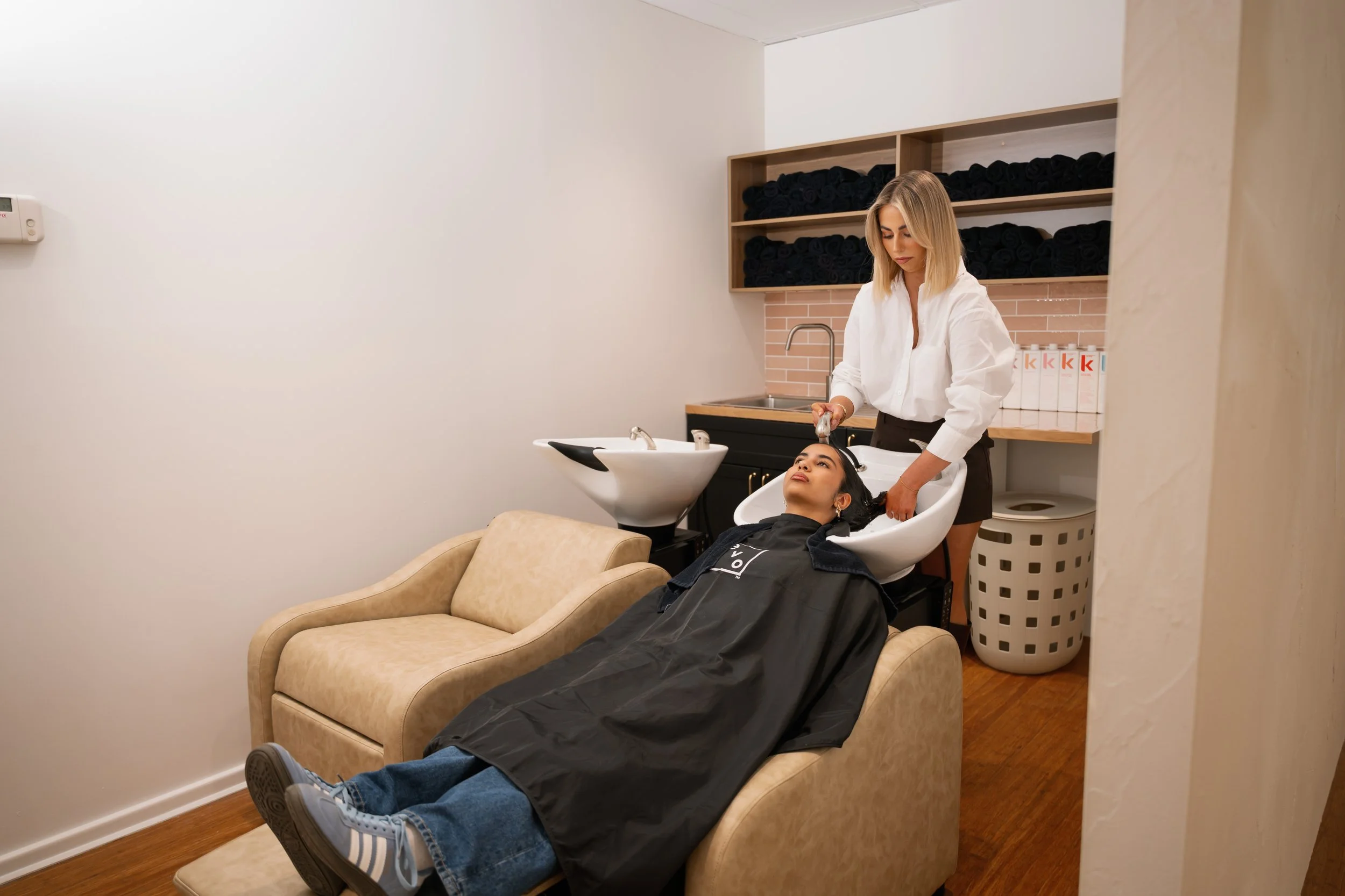 A woman receives a hair wash at a salon sink while lying back on a beige sofa, with a stylist washing her hair.