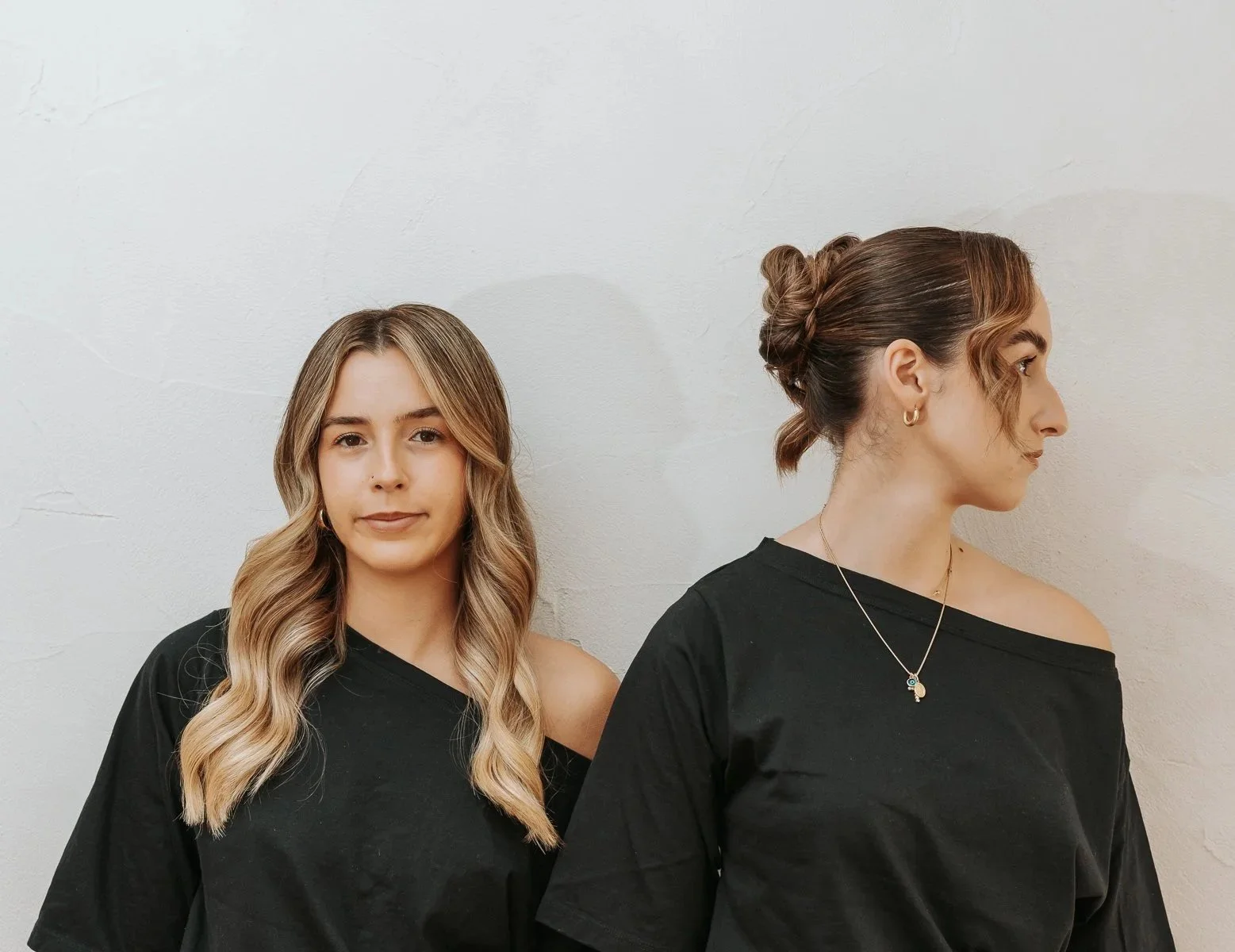 Two women wearing black tops, one facing forward with wavy blonde hair, the other in profile with brown hair styled in a bun, standing against a plain off-white wall.