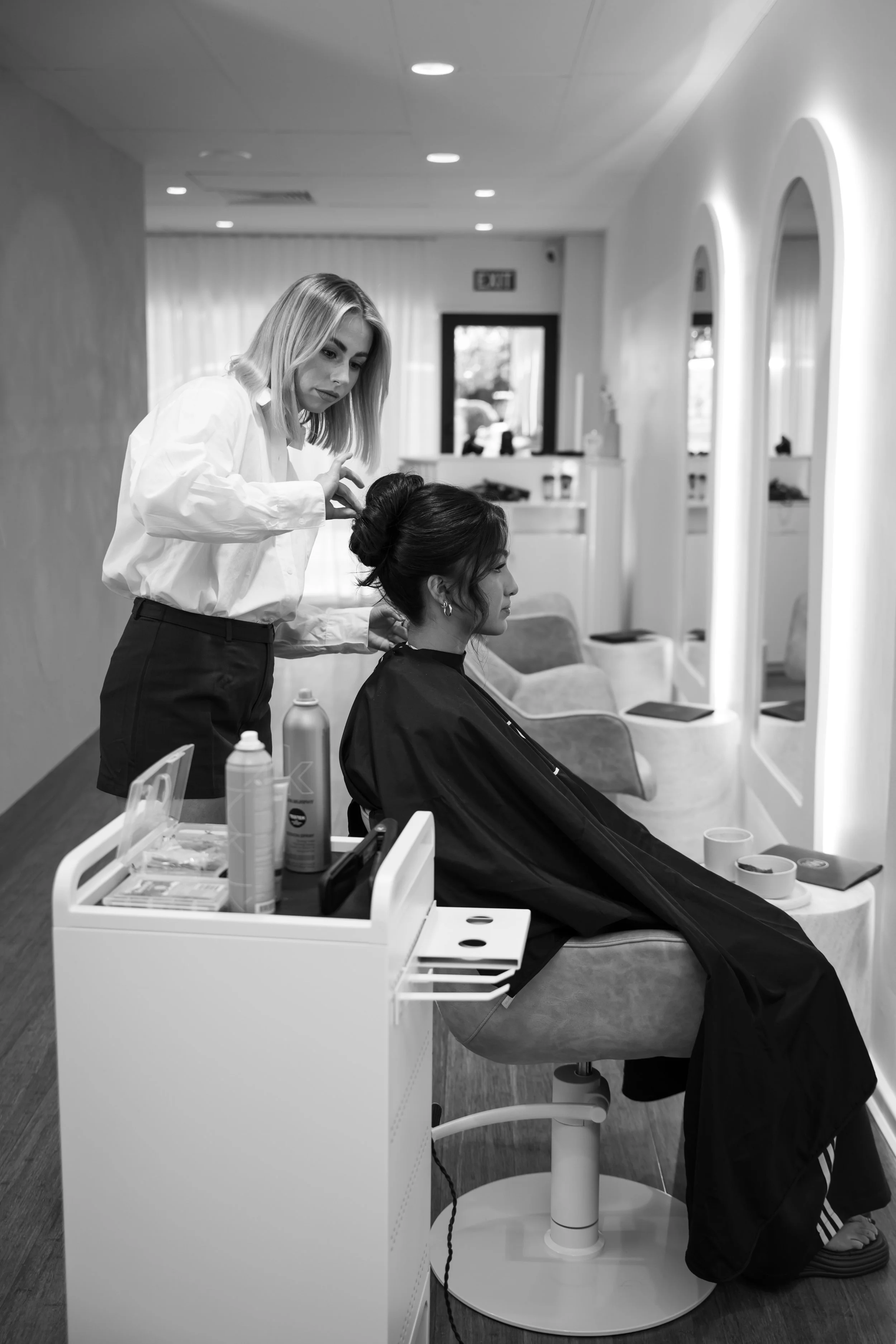A woman with dark hair is reclining in a salon chair, getting her hair styled by another woman with blonde hair, in a modern salon with white walls and large mirrors.