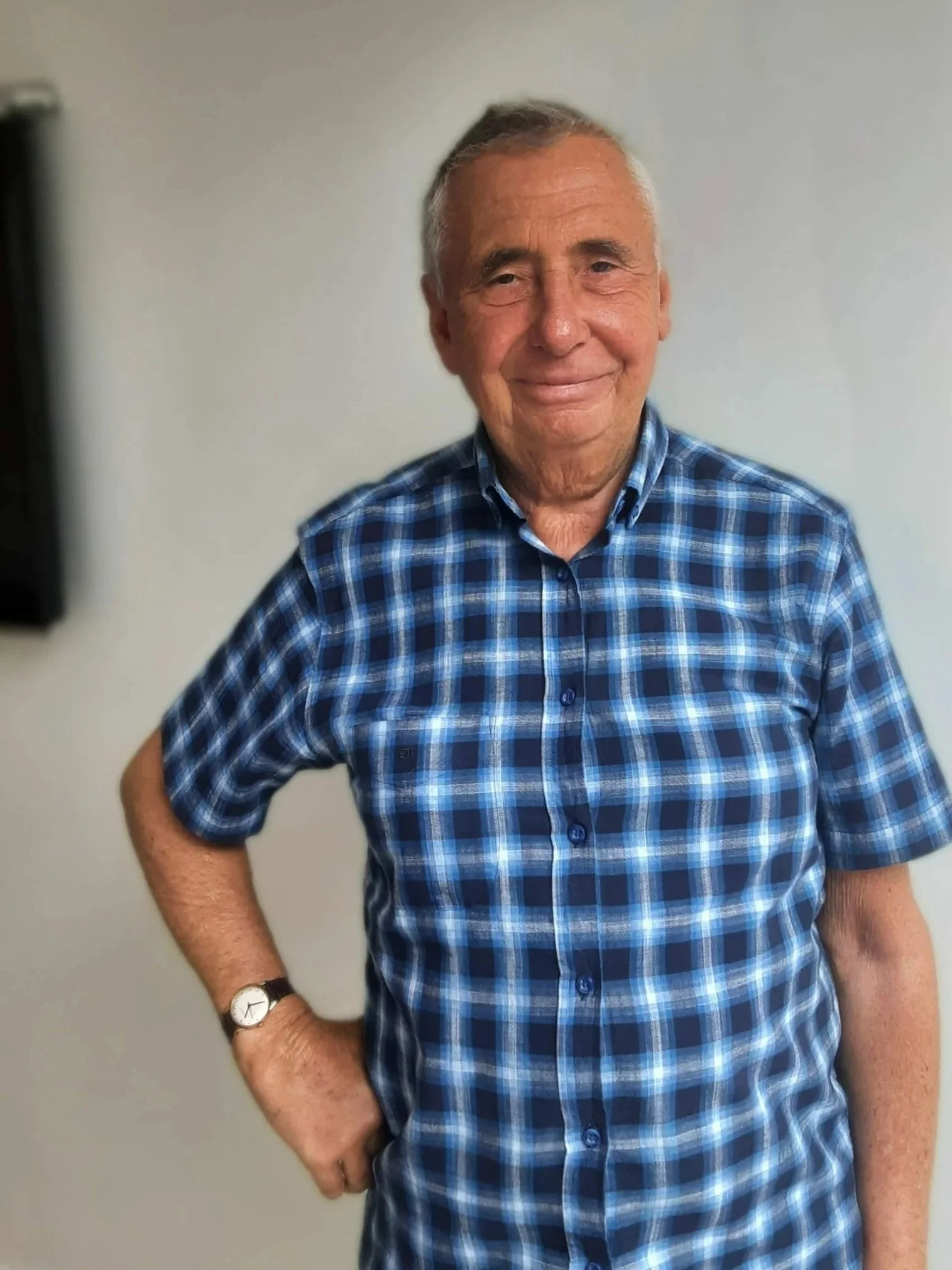 An elderly man with gray hair, wearing a blue plaid shirt, smiling, standing with one hand on his hip, against a plain light-colored wall.