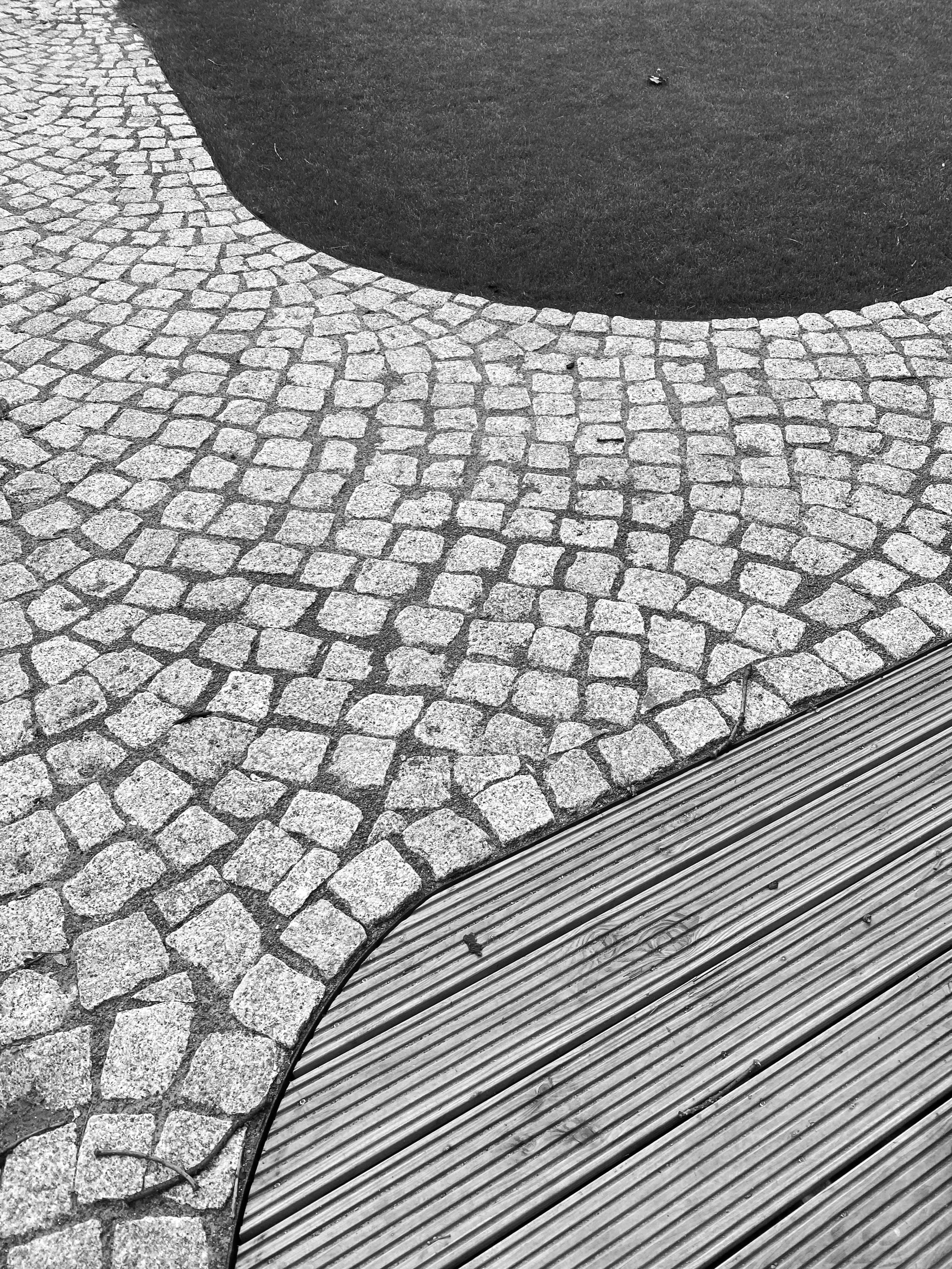 Straßenpflaster aus kleinen, quadratischen Steinen, Übergang zu glattem, schwarzen Asphalt, und darunter ein Abfluss aus Metallrohren.