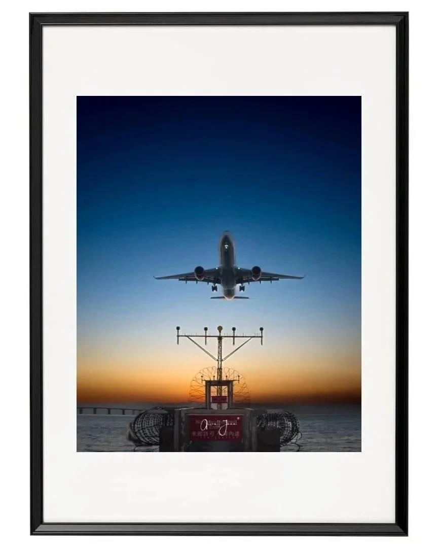 A framed aviation fine art print of a commercial airplane landing at sunset, featuring a wide white mat board and a sleek black frame.