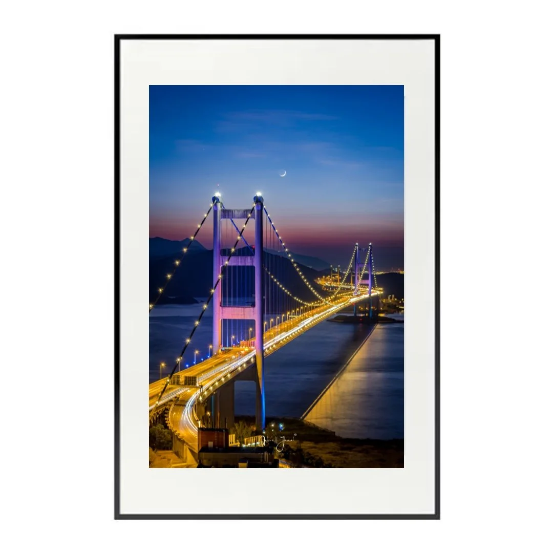 Black framed vertical photograph of a suspension bridge glowing with purple and yellow lights.