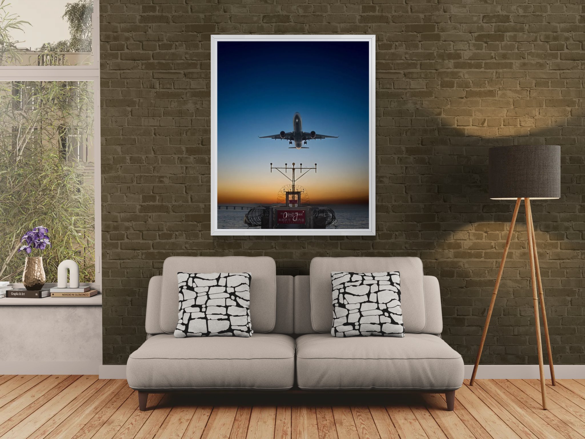 A fine art print of a commercial airplane landing at sunset, framed in white and hung on a dark brick wall above a modern grey sofa in a styled living room.