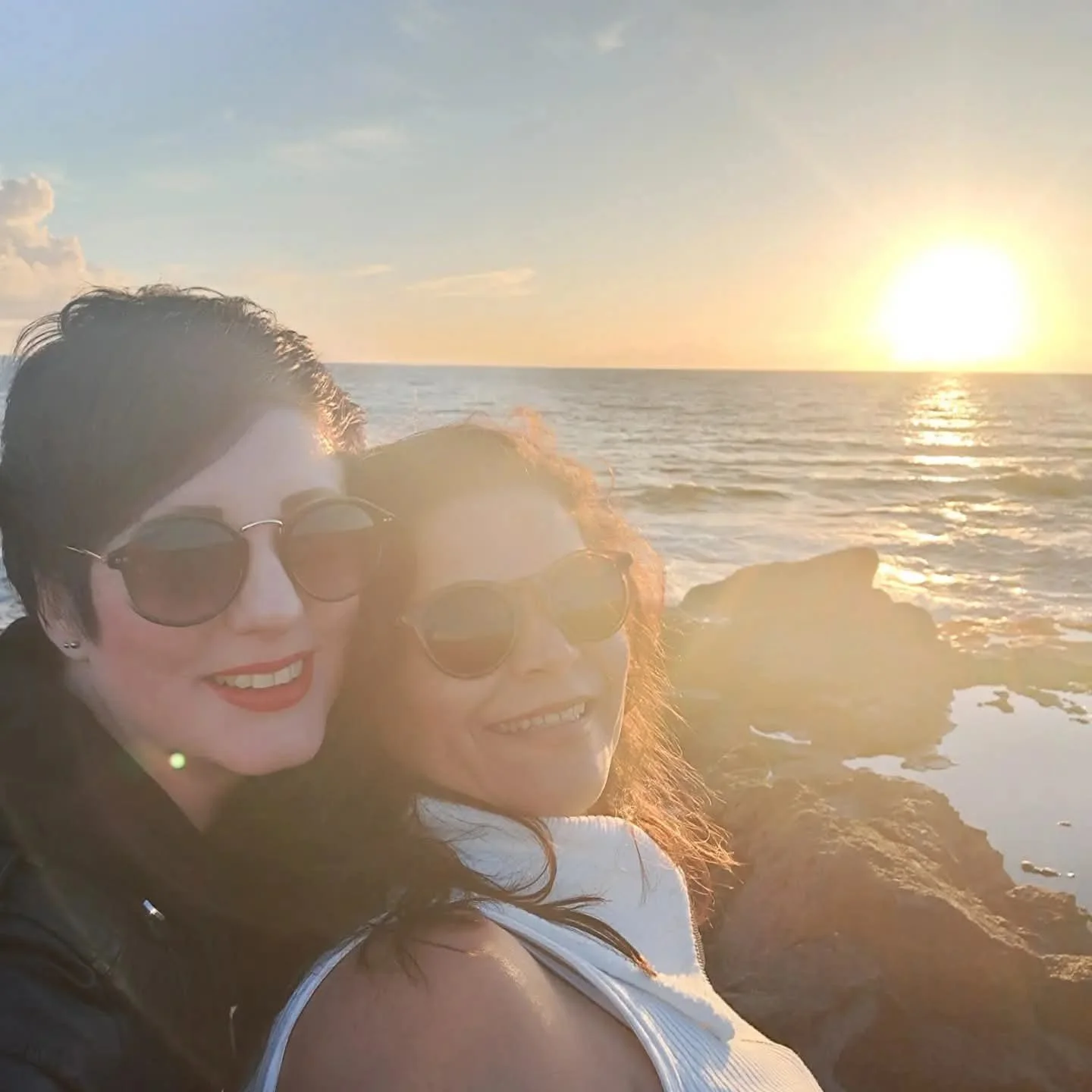 Two women with sunglasses smiling, taking a selfie at the beach during sunset with the ocean and rocks in the background.