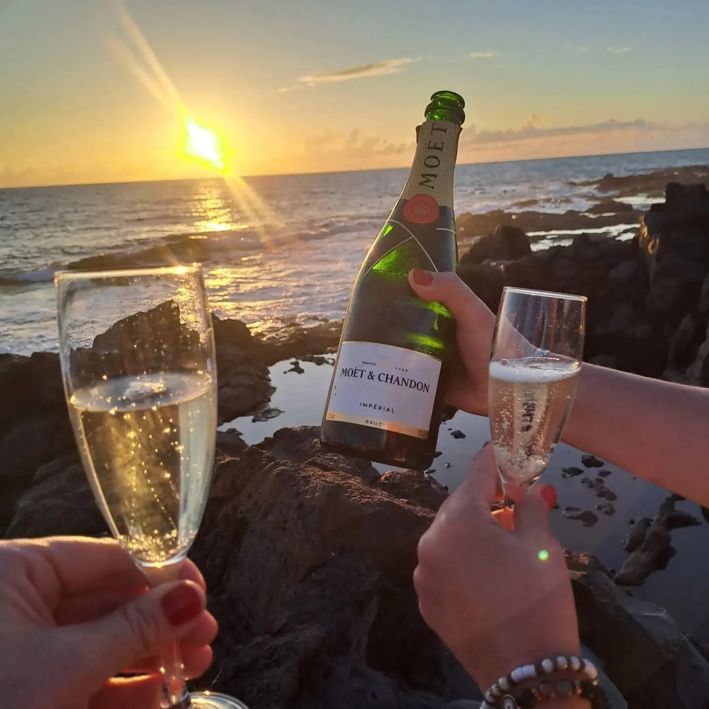 People clinking champagne glasses with a view of the setting sun over the ocean, holding a bottle of Moët & Chandon champagne in a sunset setting by the rocks.