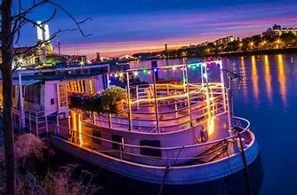 A boat decorated with multicolored lights docked on a river during sunset.