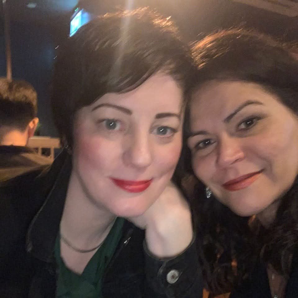 Two women with dark hair smiling at the camera in a dimly lit setting, possibly a bar or restaurant.