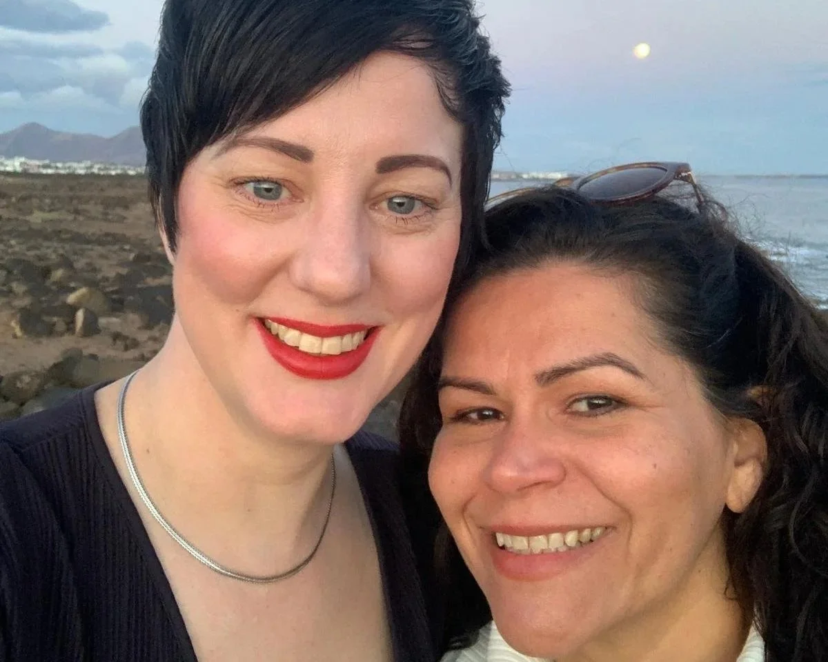 Two women smiling at the camera with a rocky beach and distant mountains in the background, likely during sunset.