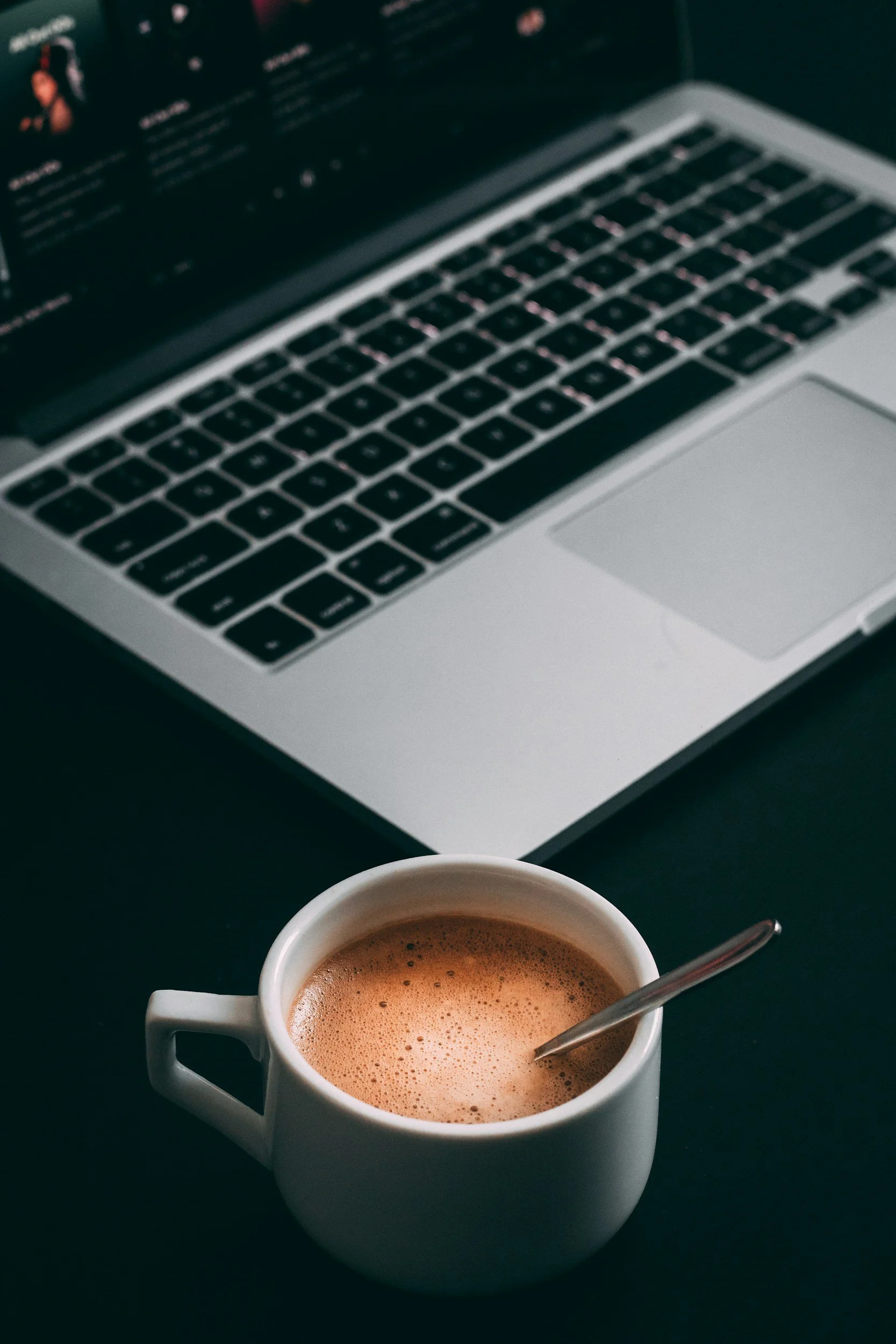 A white mug filled with hot chocolate with foam, placed on a dark surface. In the background, an open laptop with a black keyboard and a blurred screen and keyboard. A spoon is in the mug.