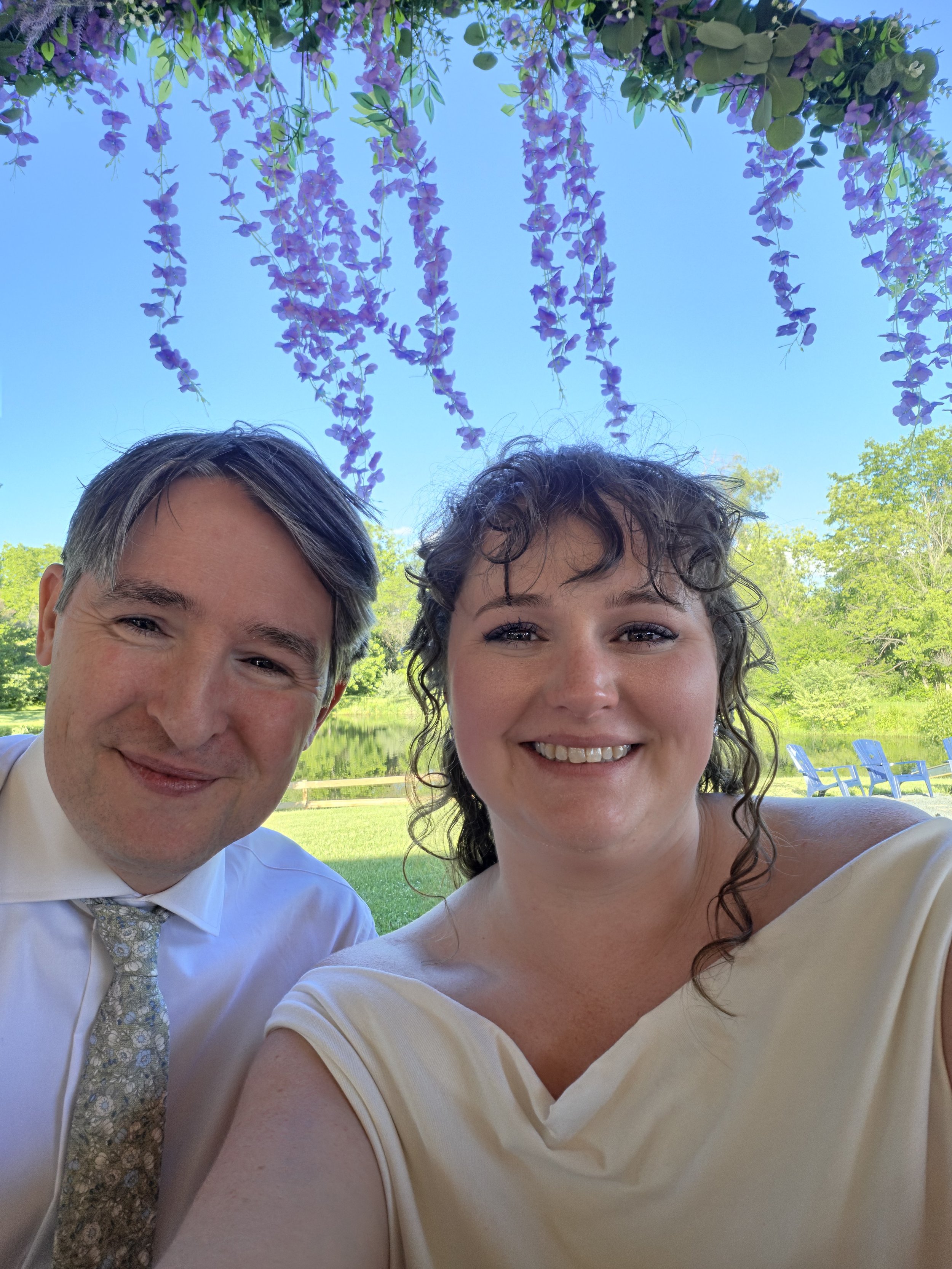 A smiling man and woman taking a selfie outdoors near a lake, with hanging purple wisteria flowers and green trees in the background on a sunny day.