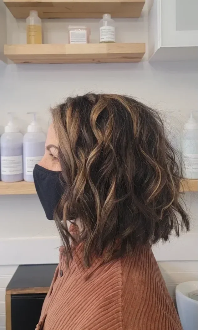 Profile of a woman with wavy, shoulder-length brown hair with blonde highlights, wearing a black face mask and a brown corduroy jacket, in a salon with shelves of hair care products behind her.
