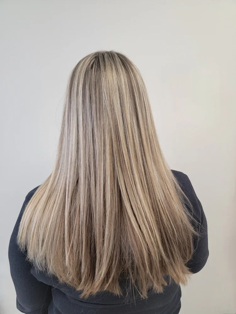 Back of a woman with long, straight, blonde hair wearing a dark top, standing against a plain light-colored wall.