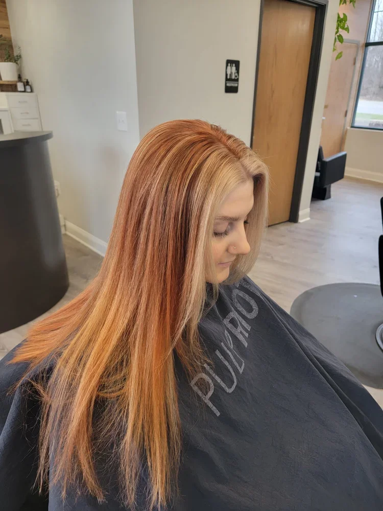 Woman with long red hair sitting in a salon chair, wearing a black cape in a hair salon.