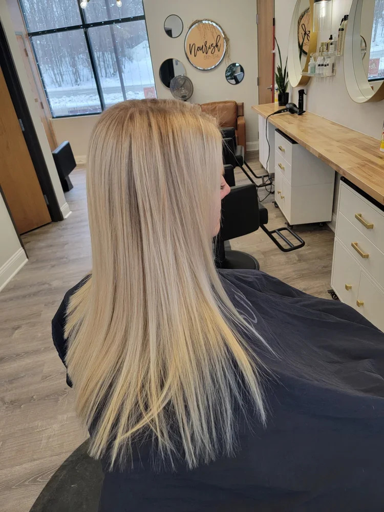 Blonde woman with straight hair sitting in a salon chair inside a hair salon, with mirrors and salon station in the background