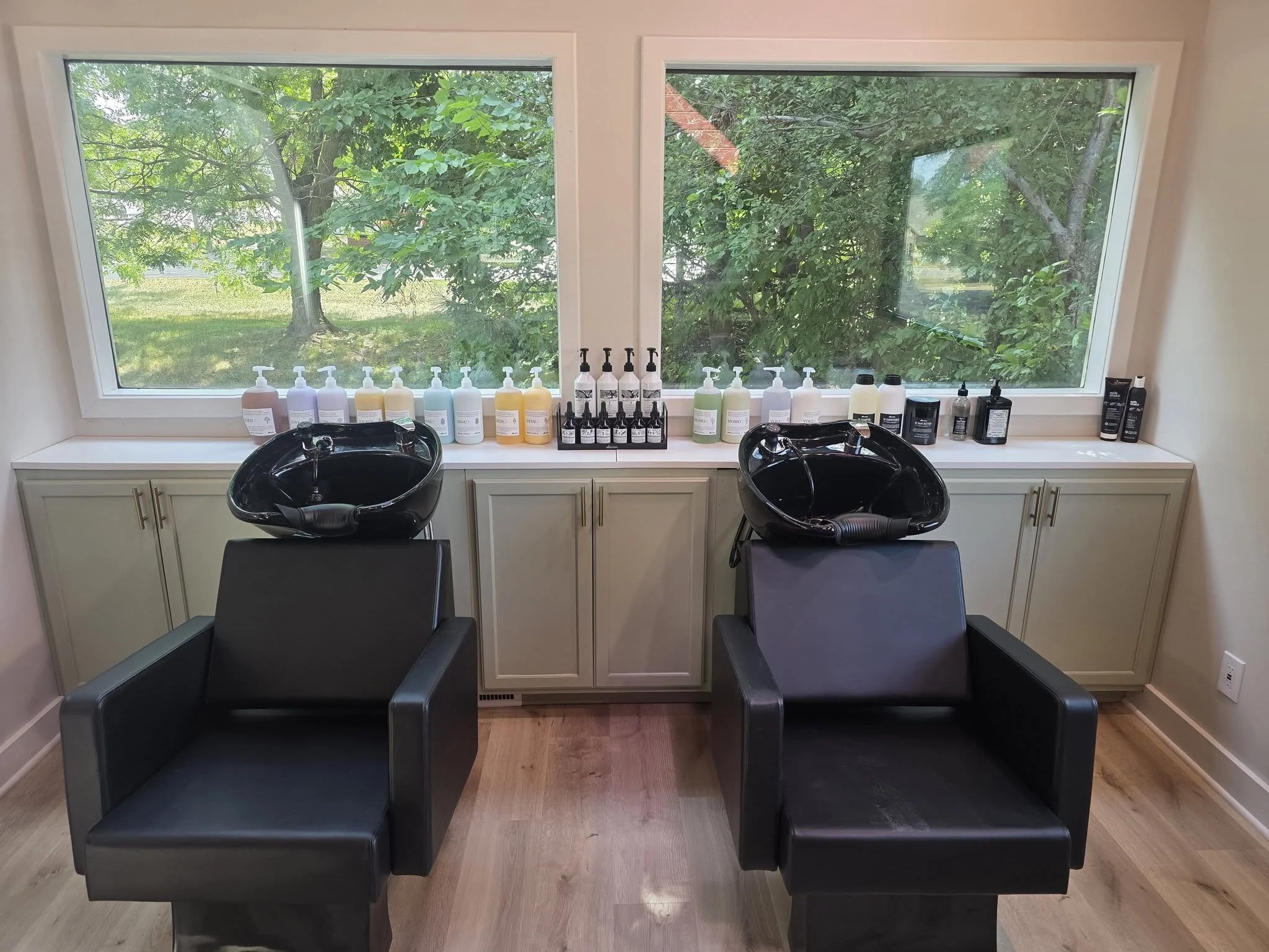 Salon with two black hair washing sinks and a row of hair care products on a white shelf in front of two large windows with a view of green trees outside.