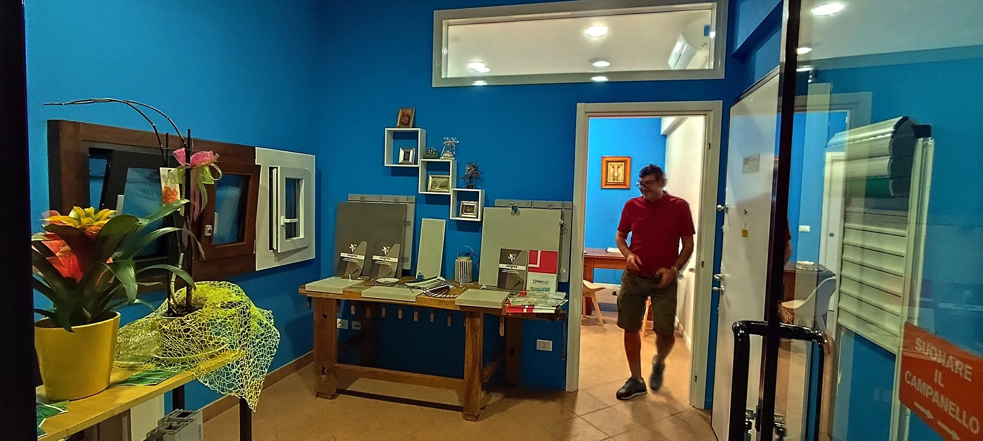 Interno di un ufficio con pareti blu, tavoli con libri e decorazioni, e una persona con pantaloncini e maglietta rossa che entra nella stanza
