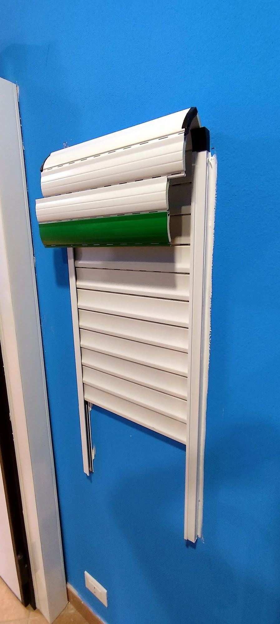 Pannello di persiana veneziana a lamelle orizzontali, bianco e verde, montato su una parete blu con presa di corrente alla base.