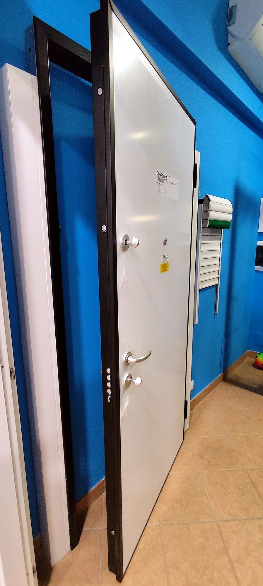 Porta blindata bianca aperta, in un ambiente con pareti blu e pavimento in ceramica, con altri elementi come pannelli e giochi colorati in una stanza.