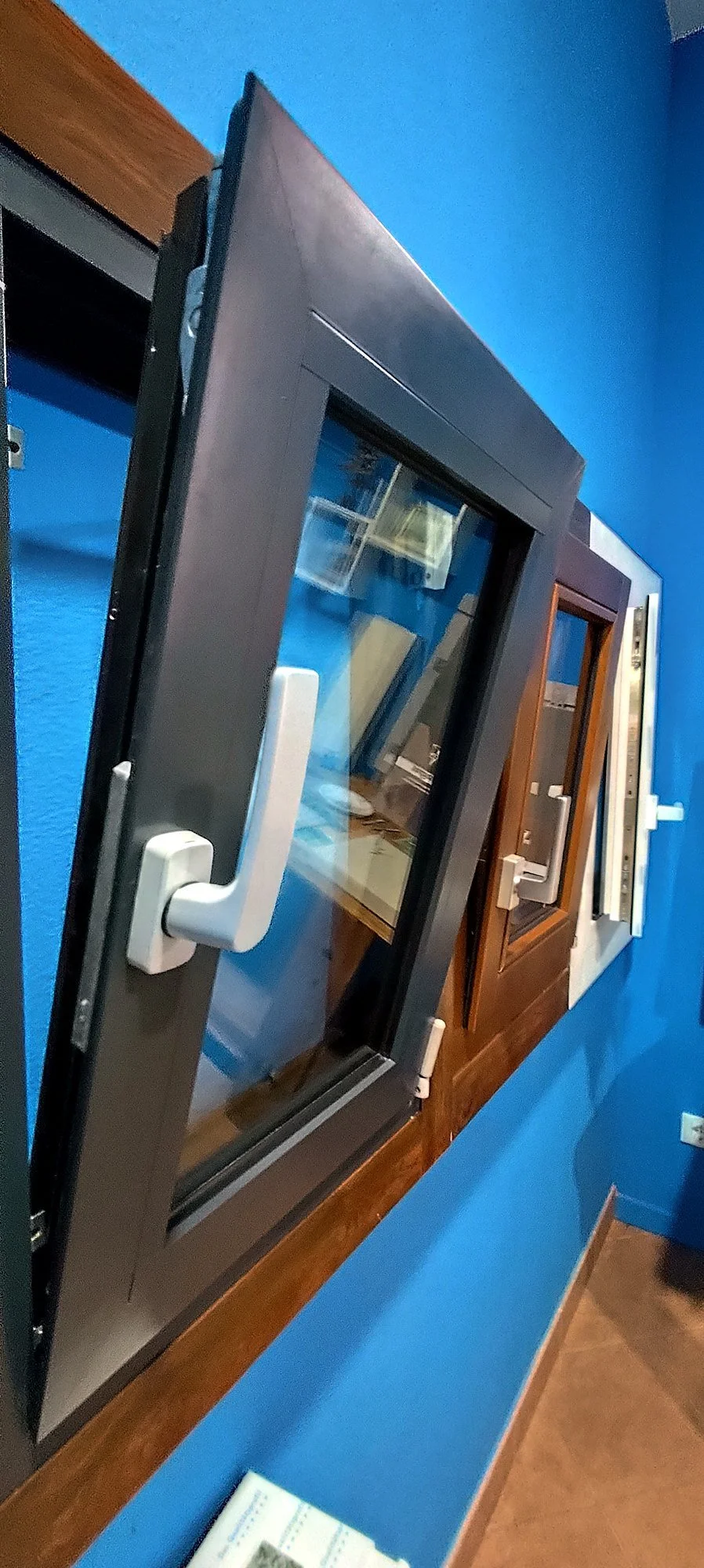 Mostra una serie di finestre con telai di vari materiali e colori, montate su una parete blu.