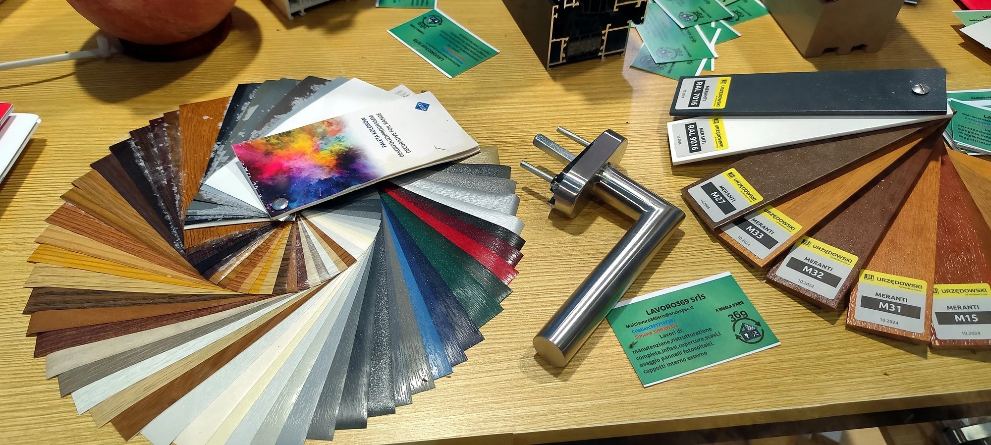 Campioni di materiali per rivestimenti e colori, tra cui colori metallici e木nuovi, con un rullo per verniciatura e vari cartoncini con codici colore e materiali. Un biglietto verde con informazioni di contatto.