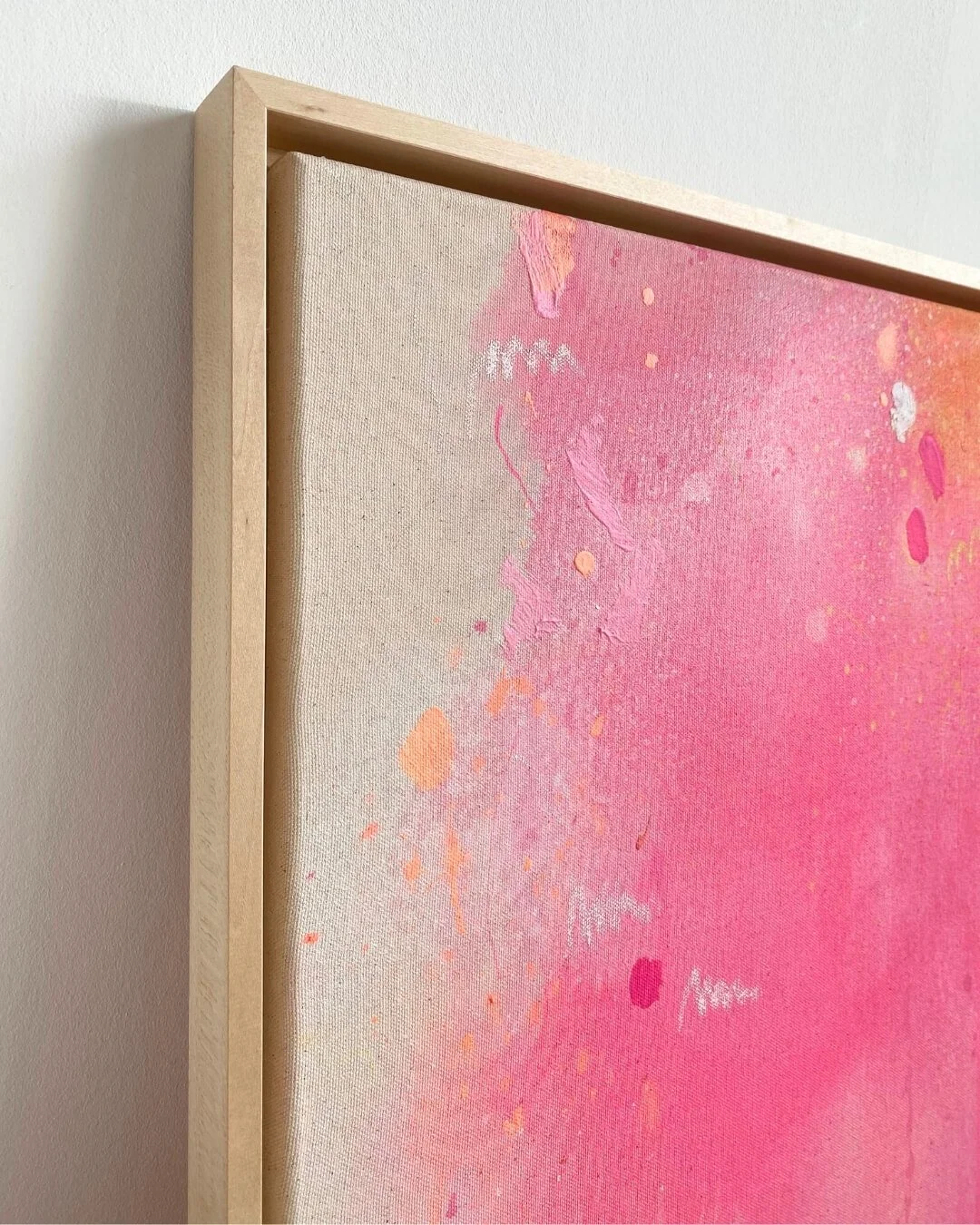 Side-angle view of a framed square abstract painting, showcasing the light wooden frame depth and the energetic pink and orange composition by Vanessa Ochi