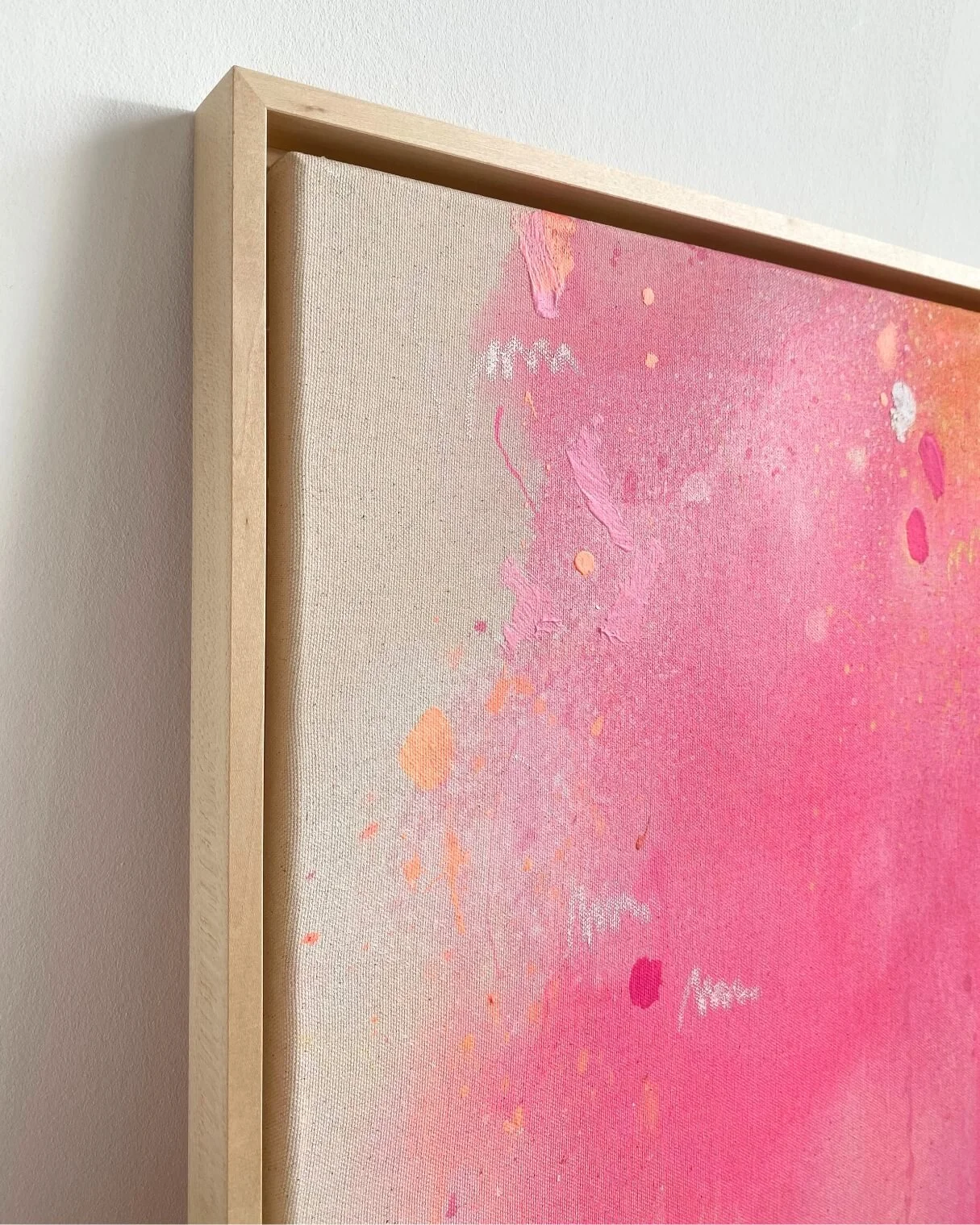 Side-angle view of a framed square abstract painting, showcasing the light wooden frame depth and the energetic pink and orange composition by Vanessa Ochi