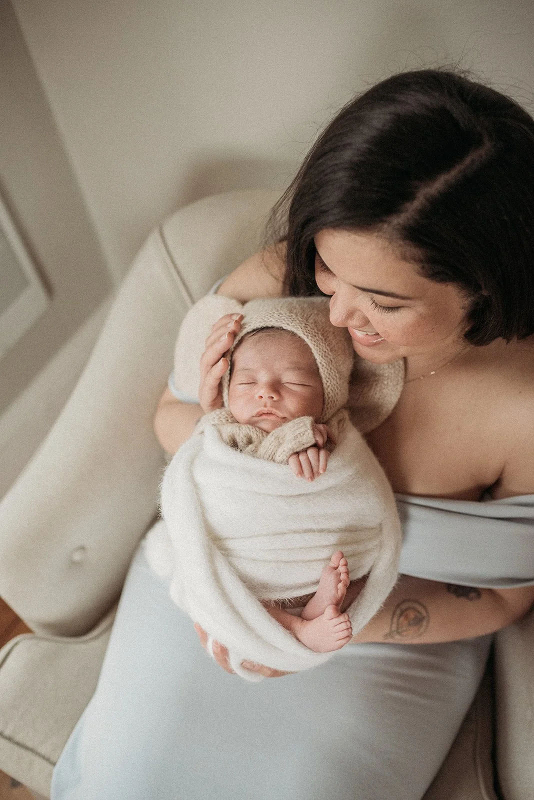 A woman holding a sleeping newborn baby swaddled in a beige blanket and wearing a knit hat and mittens, sitting on a beige bed with a light-colored background.