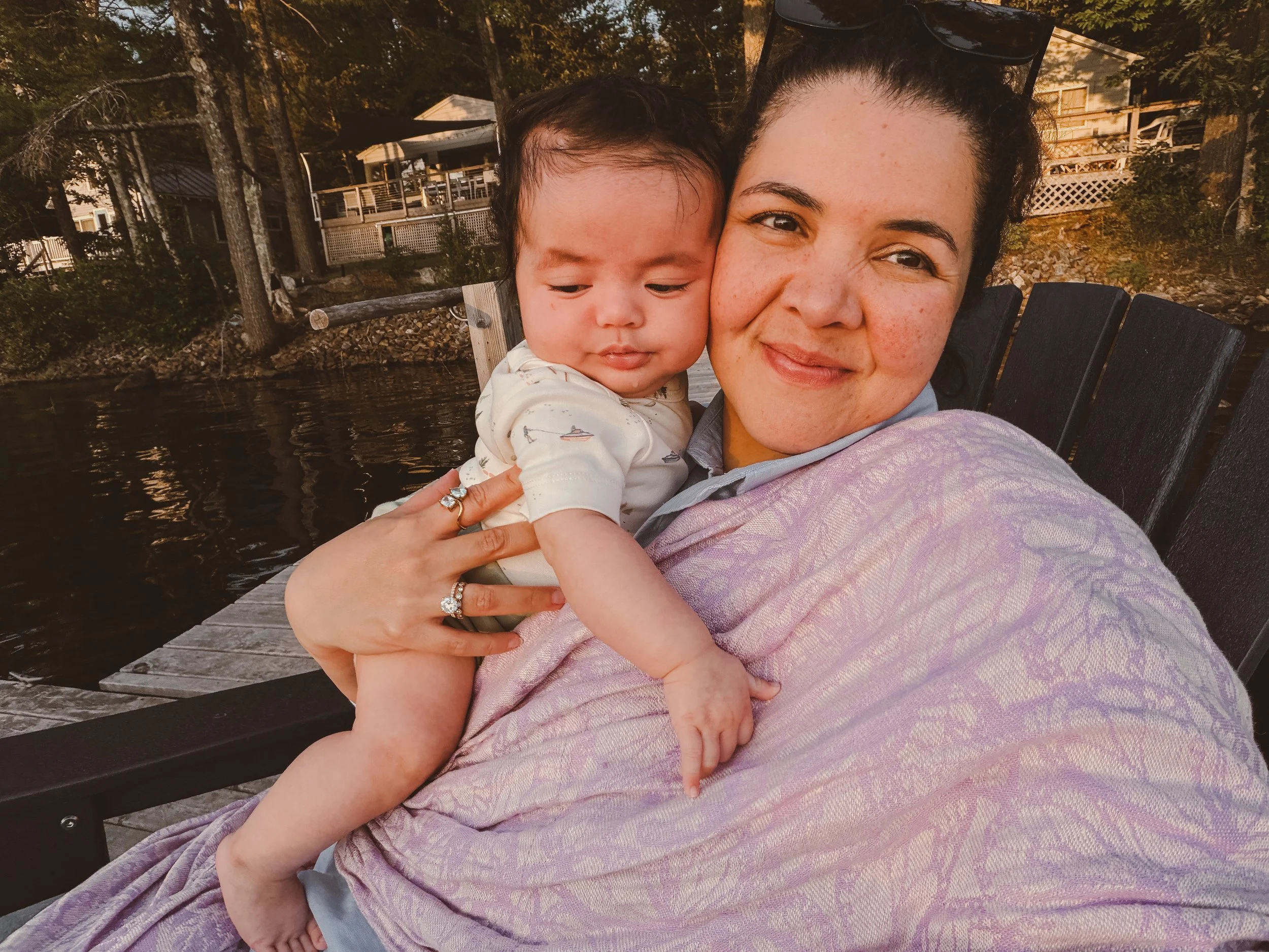 A woman holding a young child outdoors near water with trees and houses in the background during sunset.