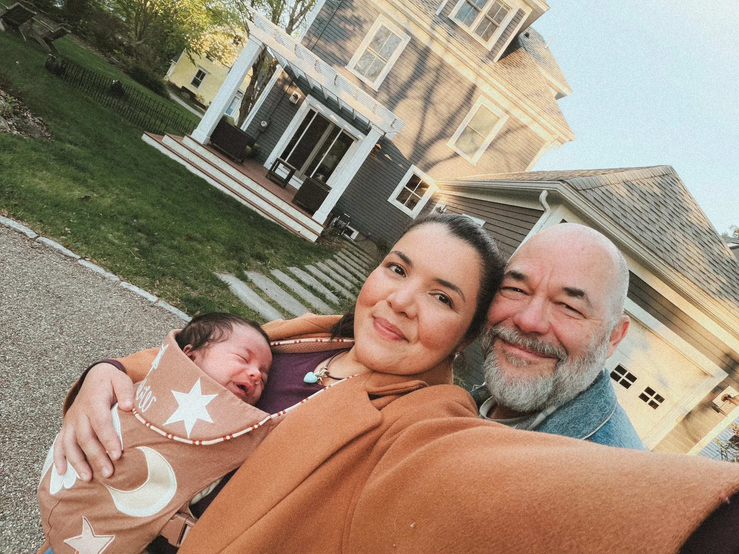 A family of four, including a woman, a man, and two babies, standing outside near a house with a lawn and trees in the background in the late afternoon or early evening.