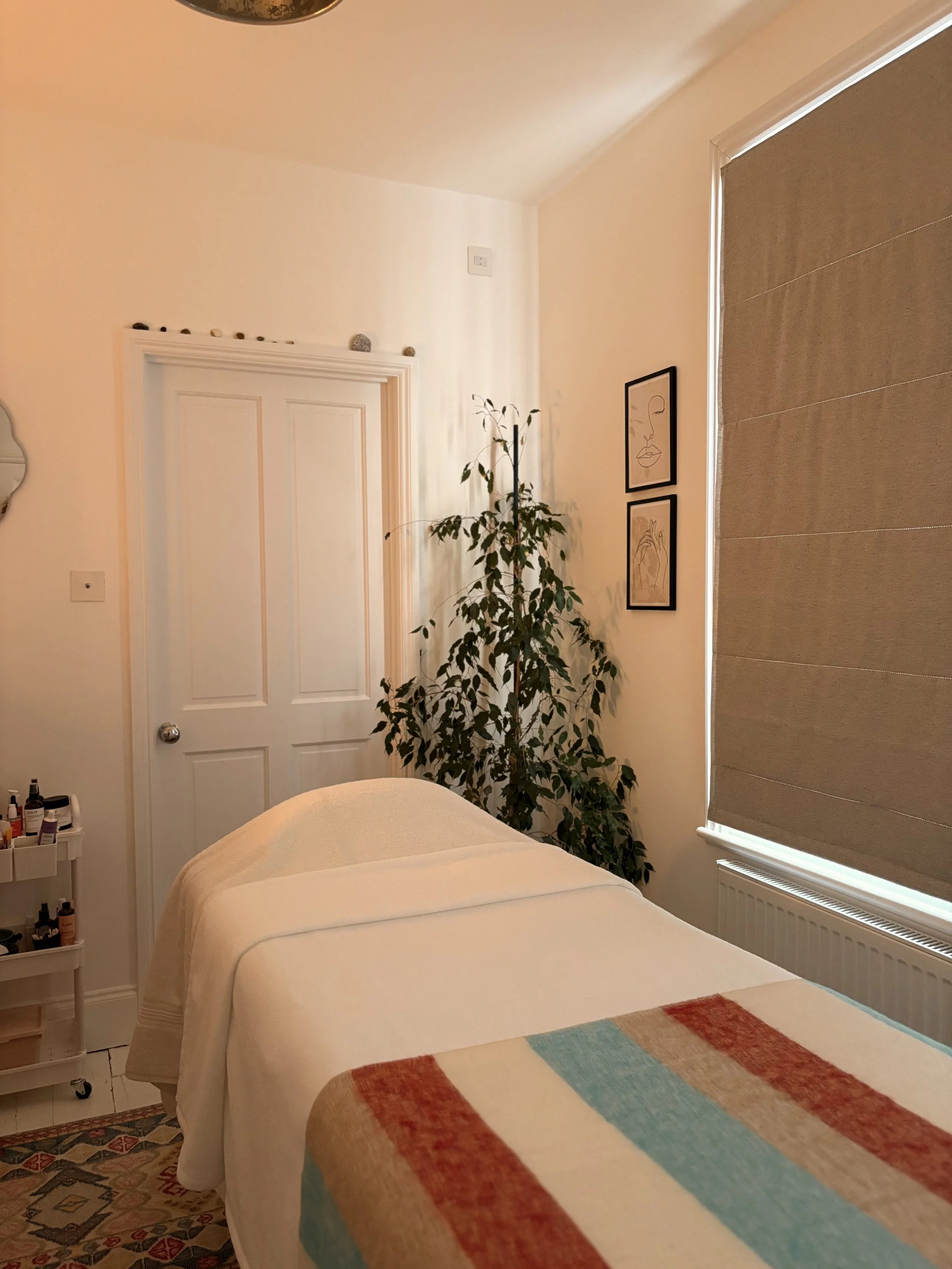 A cozy massage room with a bed covered in a beige sheet and a colorful striped blanket. There is a gray roll of towel on the bed. A white rolling cart with skincare products is beside the bed. A large leafy plant is in the corner, and two line art fr