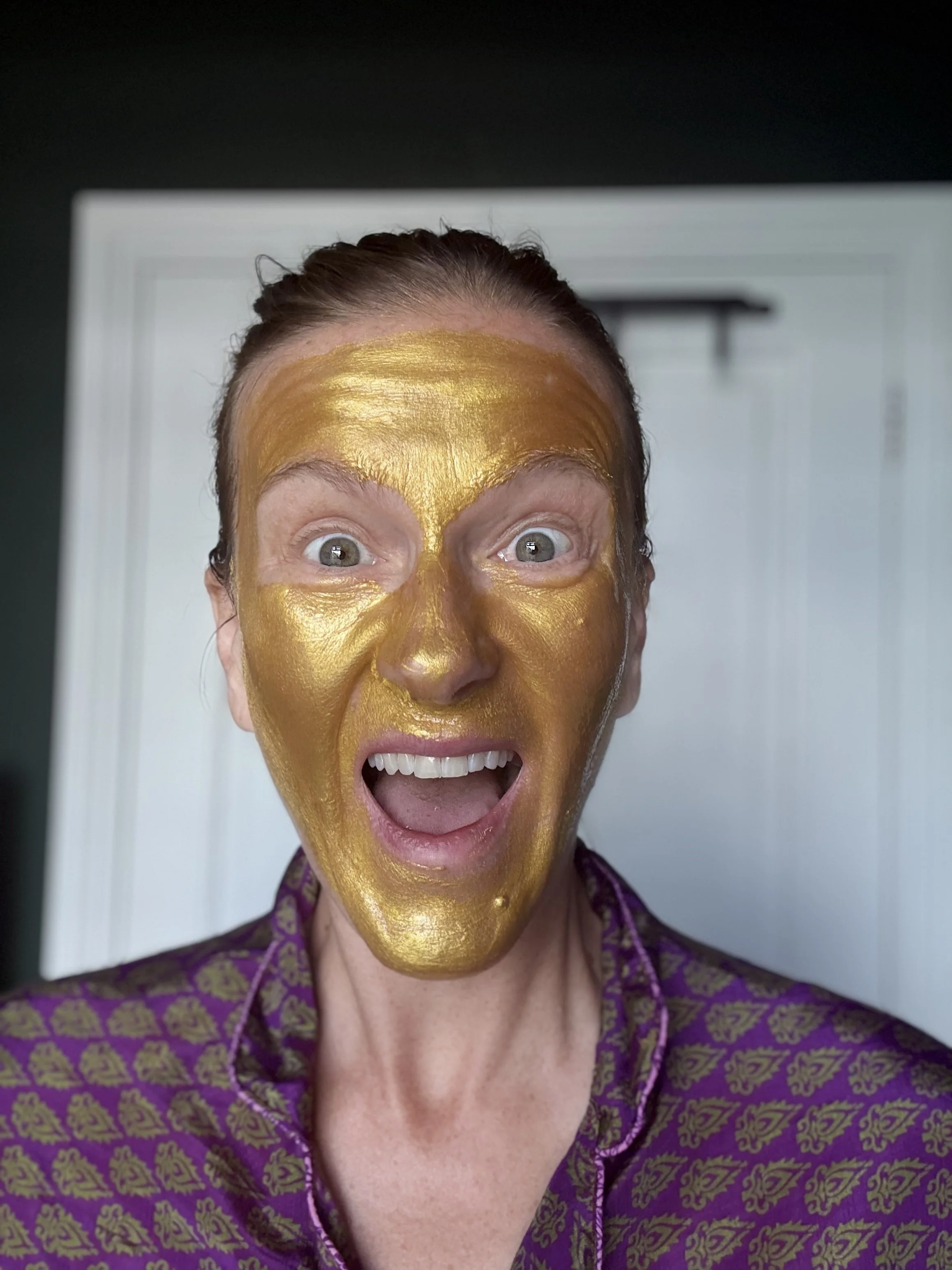 A woman with a gold facial mask covering her face, open mouth, and wide eyes, wearing a purple patterned shirt.