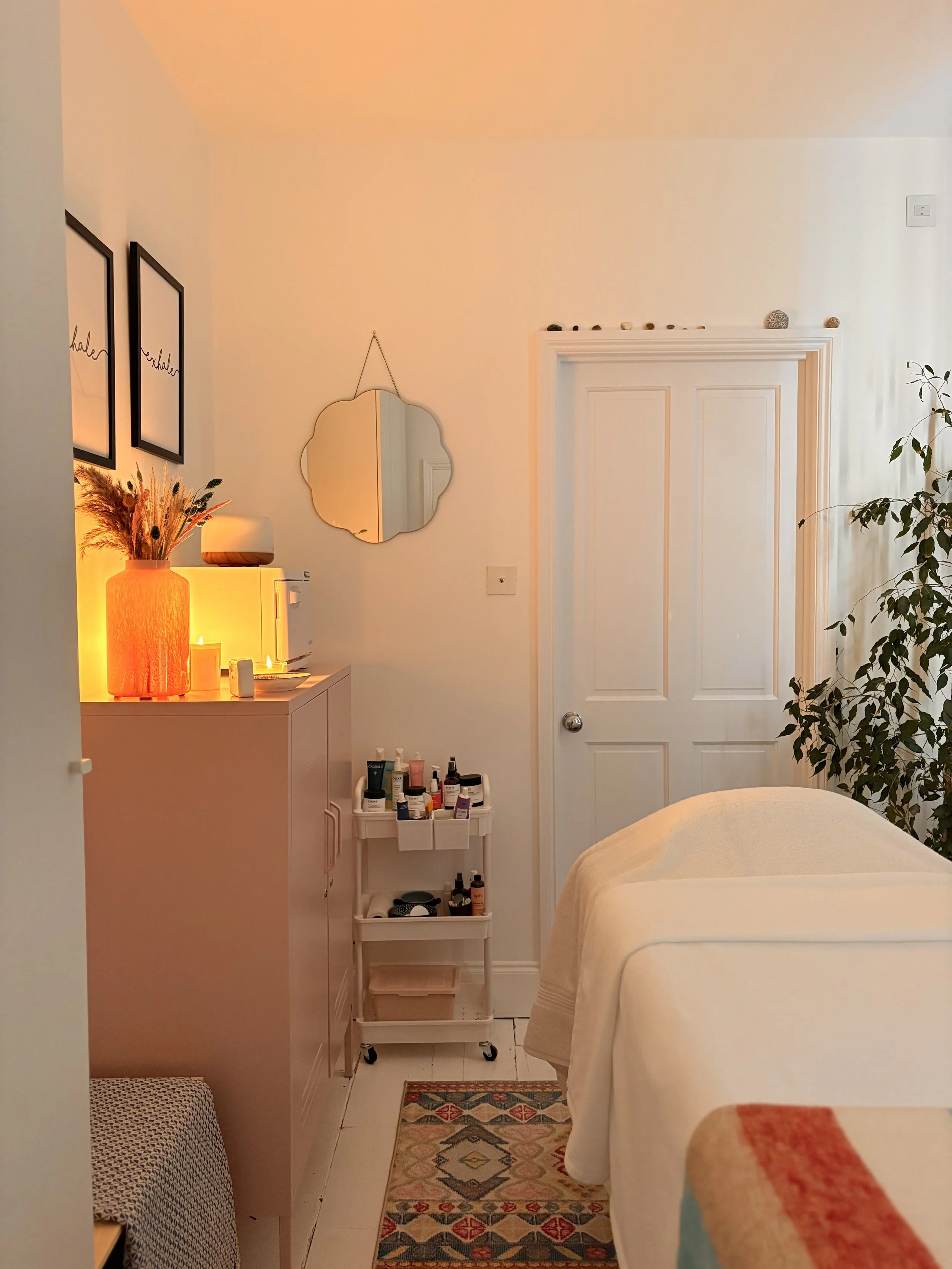 A cozy bedroom with a white bed, a white cabinet with toiletries, decorative plants, a wall mirror, and two framed quotes that say 'breathe' and 'inhale' on the wall. Warm lighting and a colorful rug complete the space.