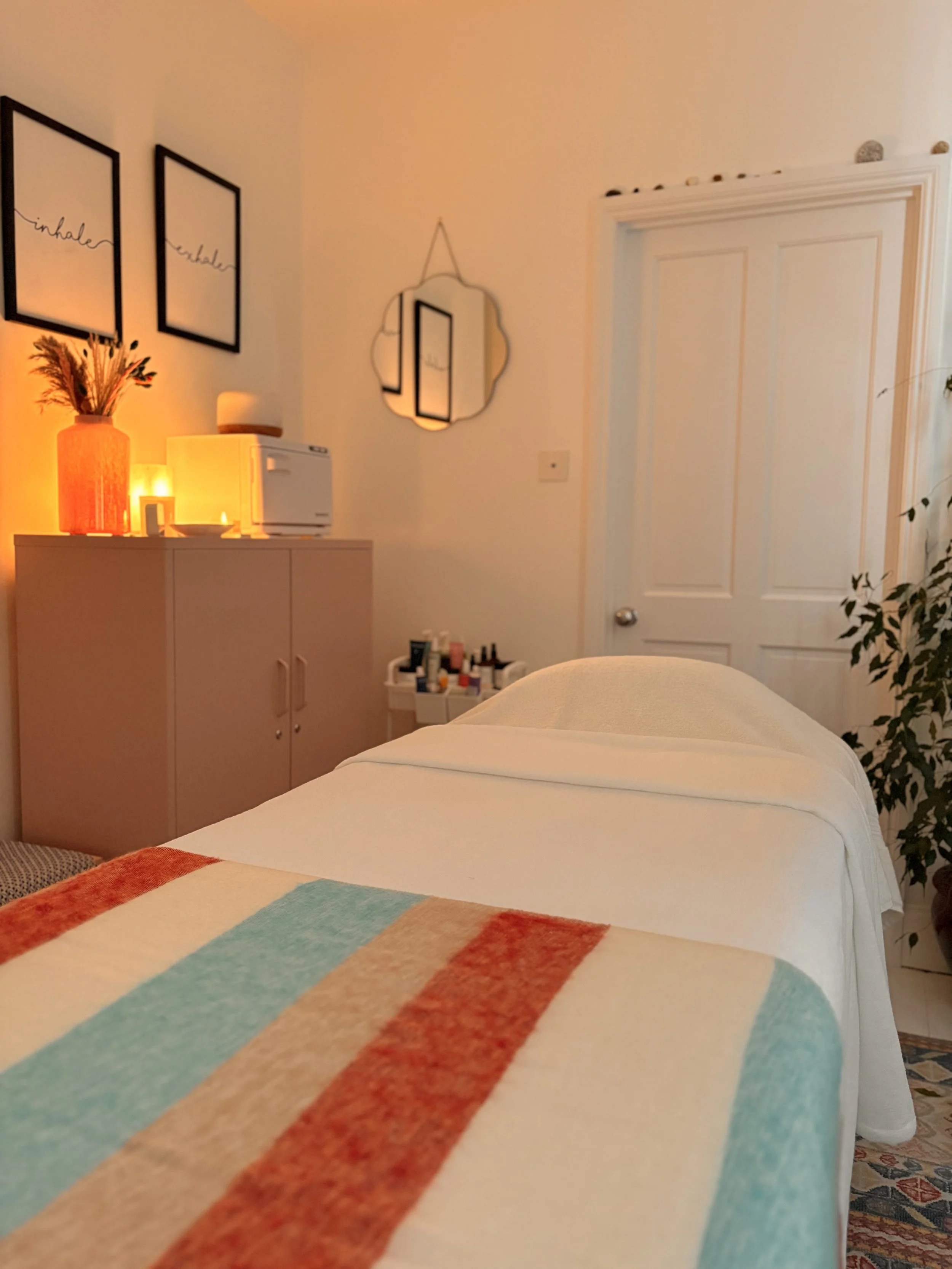 A cozy bedroom with a bed, colorful striped blanket in the foreground, a dresser with a vase of dried flowers, a small lamp, and skincare products on top. Two framed wall art pieces say 'inhale' and 'exhale'. A decorative mirror and a door are visibl