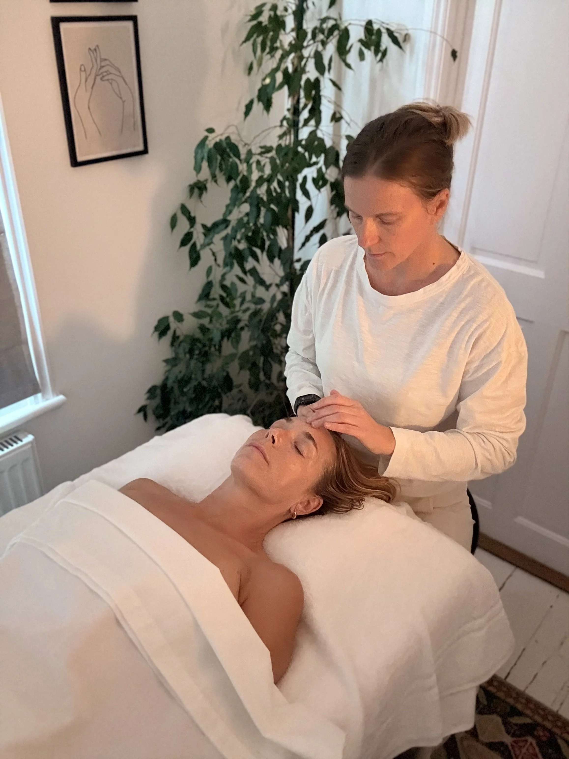 A woman lying face up on a massage table with her eyes closed, receiving a facial massage or skincare treatment from a woman standing beside her in a cozy room.