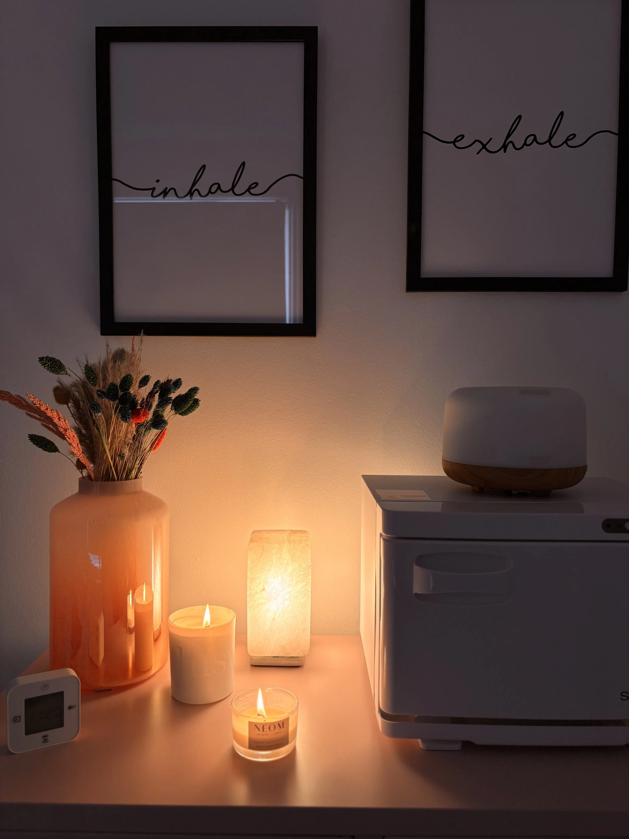 A table with illuminated candles, a vase with dried flowers, framed wall art with the words 'inhale' and 'exhale', a small device displaying temperature, and a small appliance.
