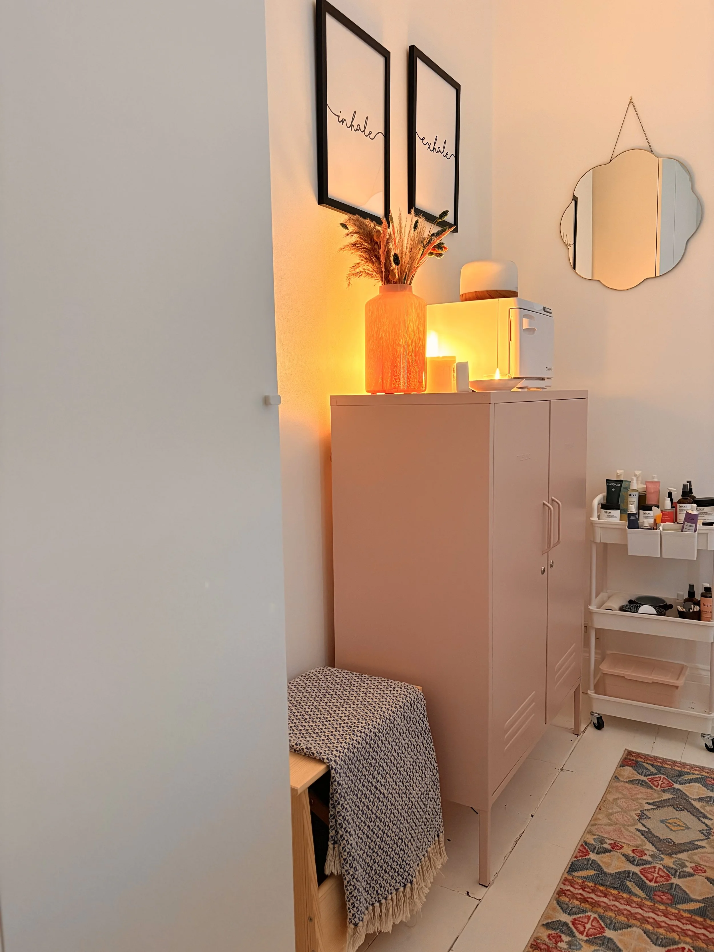 A cozy corner in a room with a pink cabinet, a framed wall art with the word 'inhale' repeated, a large orange vase with dried flowers, a small ottoman with a patterned throw, a mirror hanging on the wall, a white cart with skincare products, a small