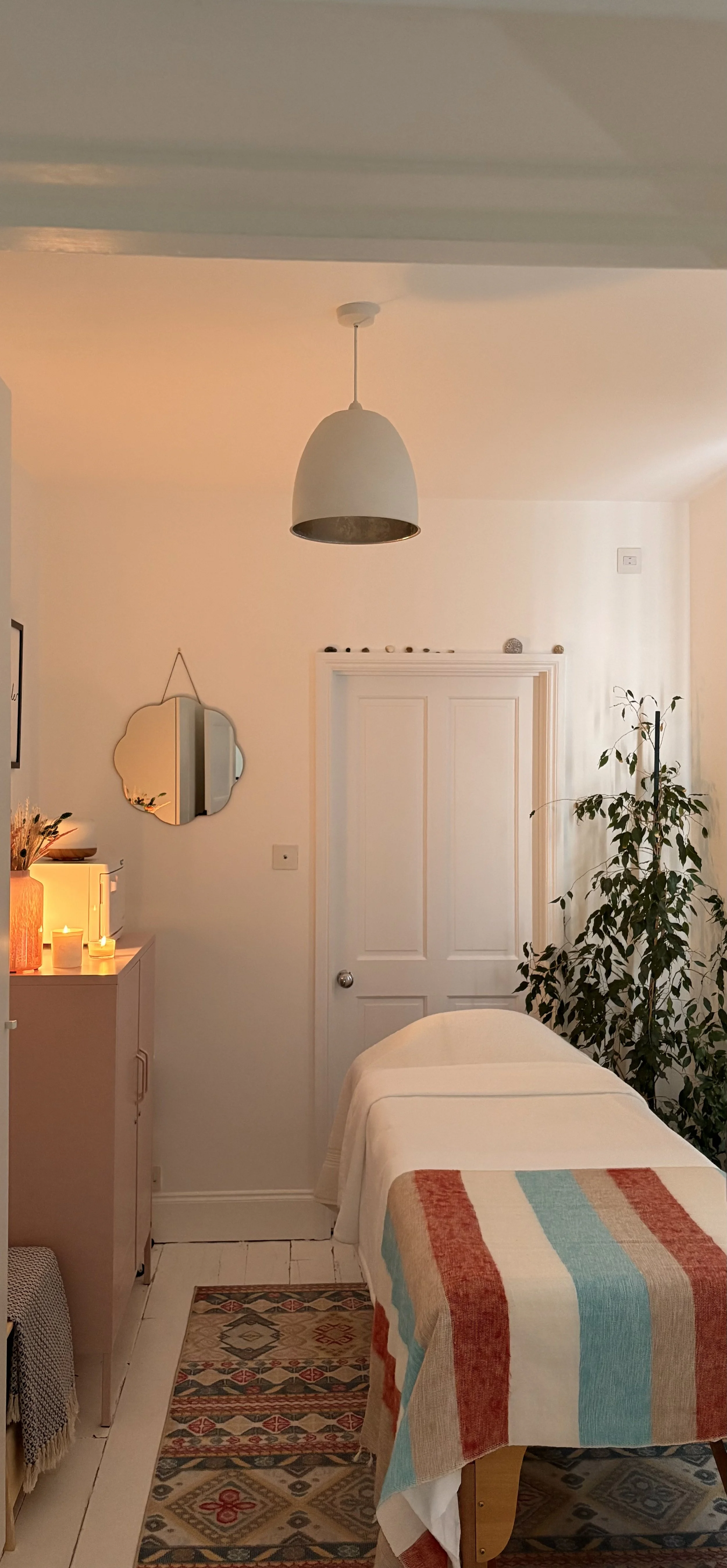 A cozy bedroom with a bed covered in a striped blanket, a white door, a potted plant, a mirror, and candles on a small cabinet, with warm lighting and decorative accents.