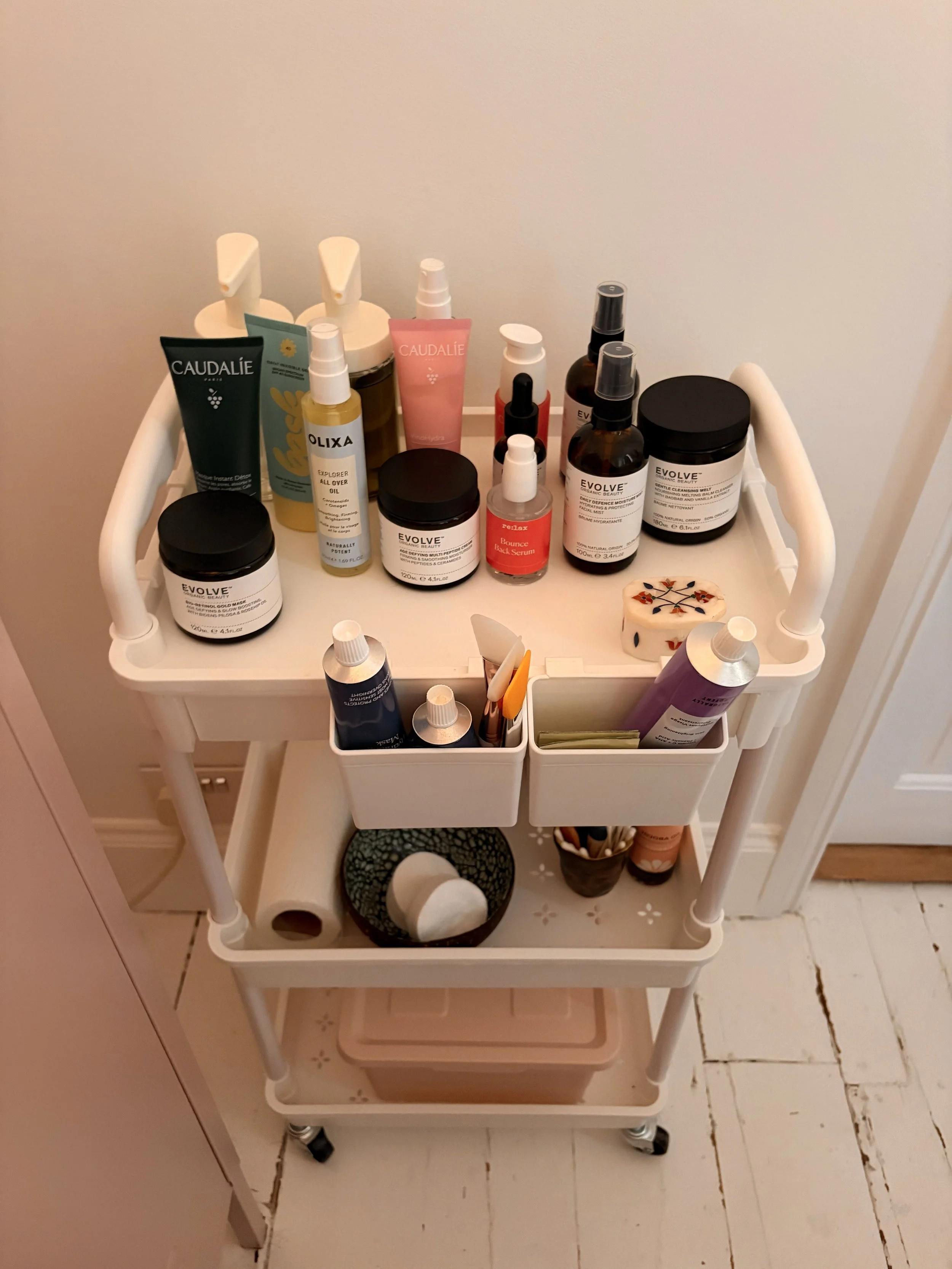 A white rolling cart with two shelves, holding various skincare and cosmetic products on the top shelf, including bottles, jars, and tubes, with some containers labeled 'Evolve' and 'Caudalie'. The bottom shelf contains more bottles, a small plant, a