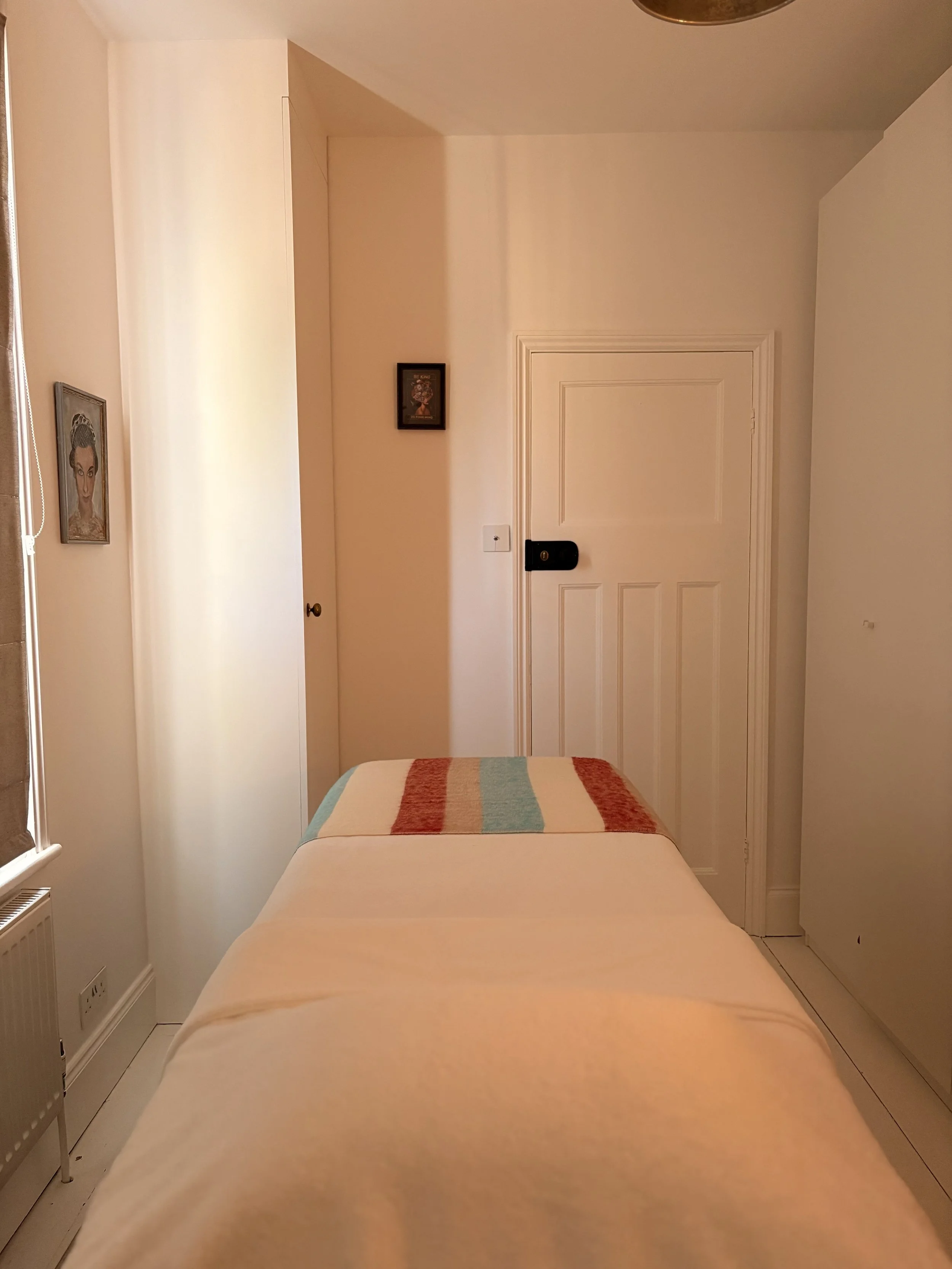 A cozy bedroom with a bed, colorful striped blanket, white walls, framed pictures, a closed door, and a white radiator.