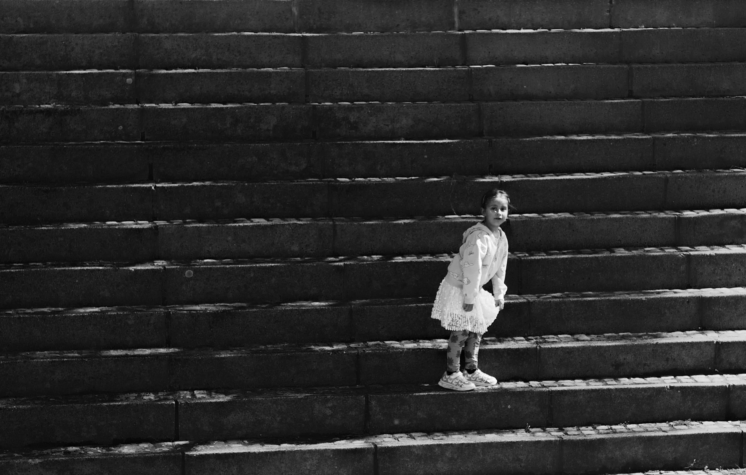 A young girl standing on steps, looking at the camera. The image is in black and white, and the girl is dressed in a light-colored jacket, patterned leggings, a skirt, and sneakers.