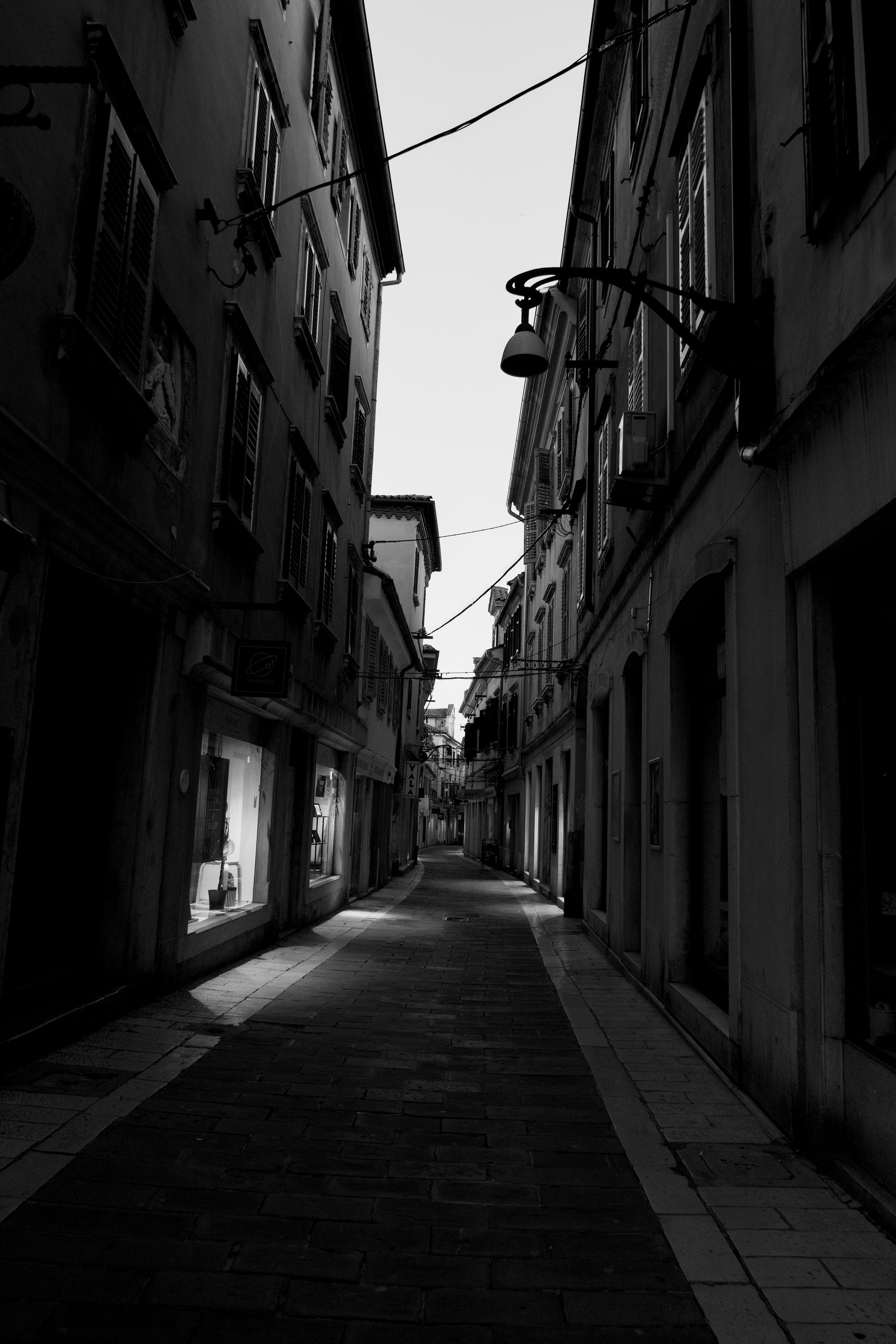 A narrow, empty street in an old European town, lined with tall buildings on both sides, with windows and shutters, some shops with display windows, and a few hanging lights above. It appears to be late afternoon or early evening.