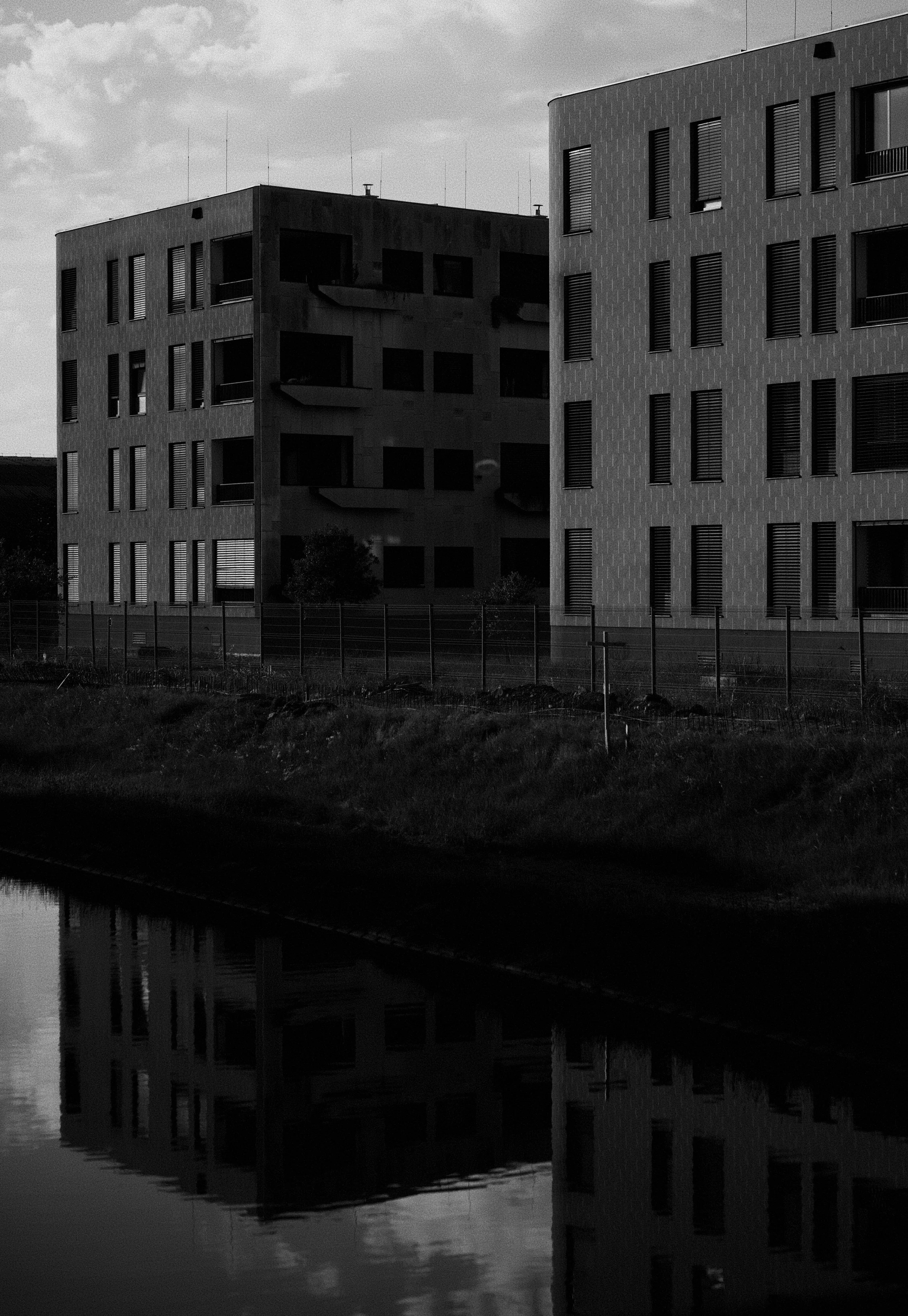 Black and white photo of modern multi-story buildings with rectangular windows and balconies, reflecting on a small body of water in the foreground.
