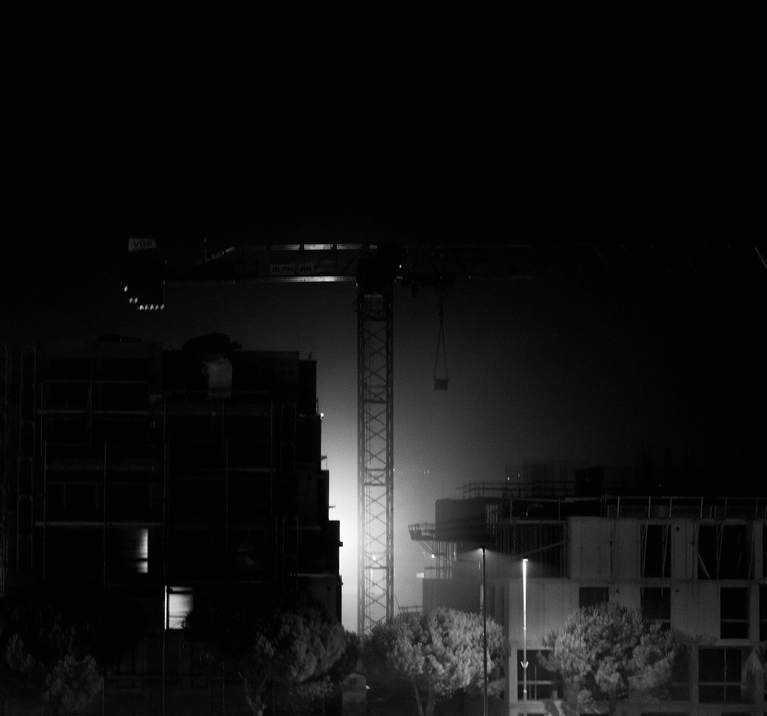 A dark cityscape or construction site at night with silhouettes of buildings and a tall electrical tower or crane, some illuminated signs on structures, and minimal lighting.