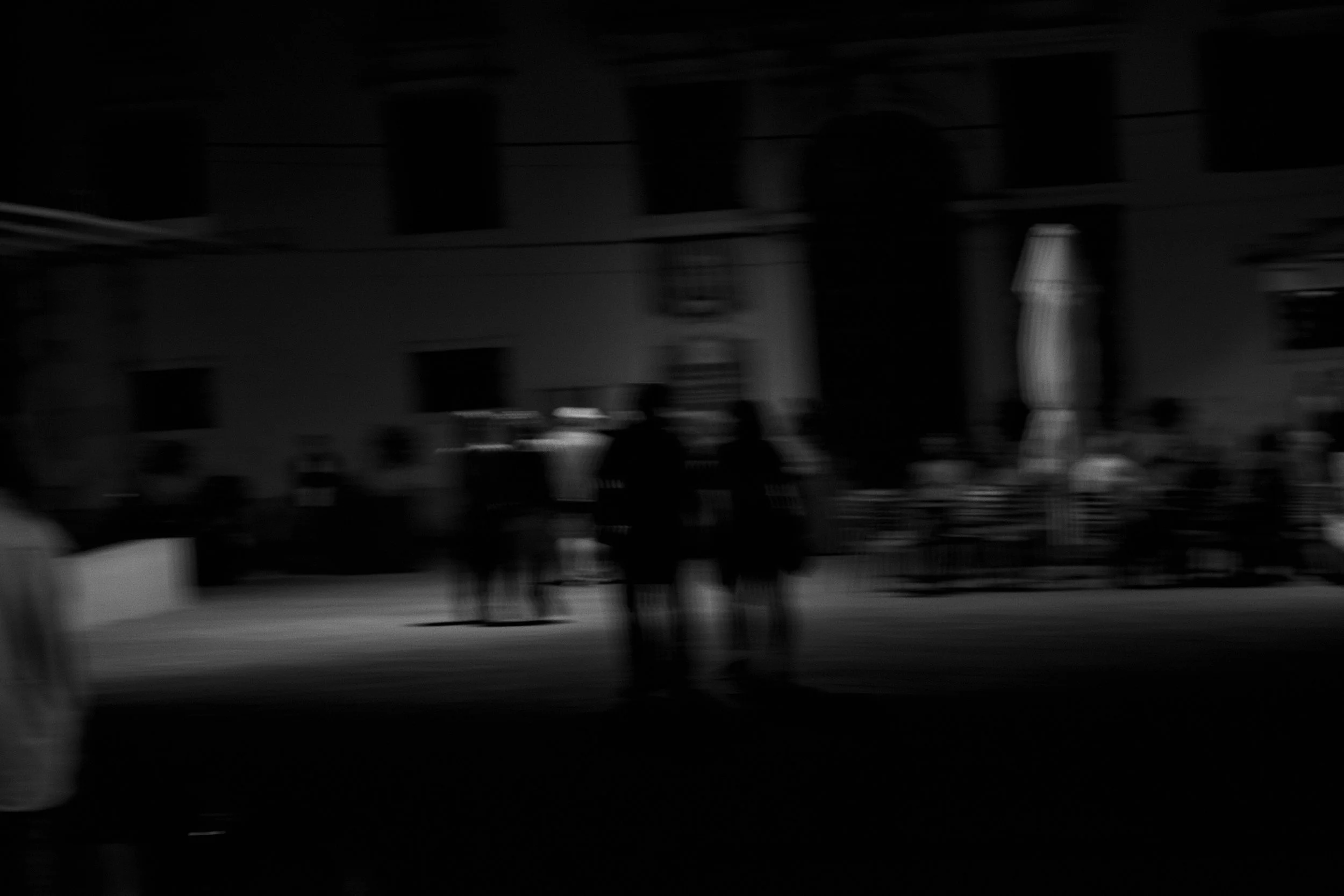 Black and white photograph of a building facade with windows, two silhouettes of people standing in front, and a street scene.