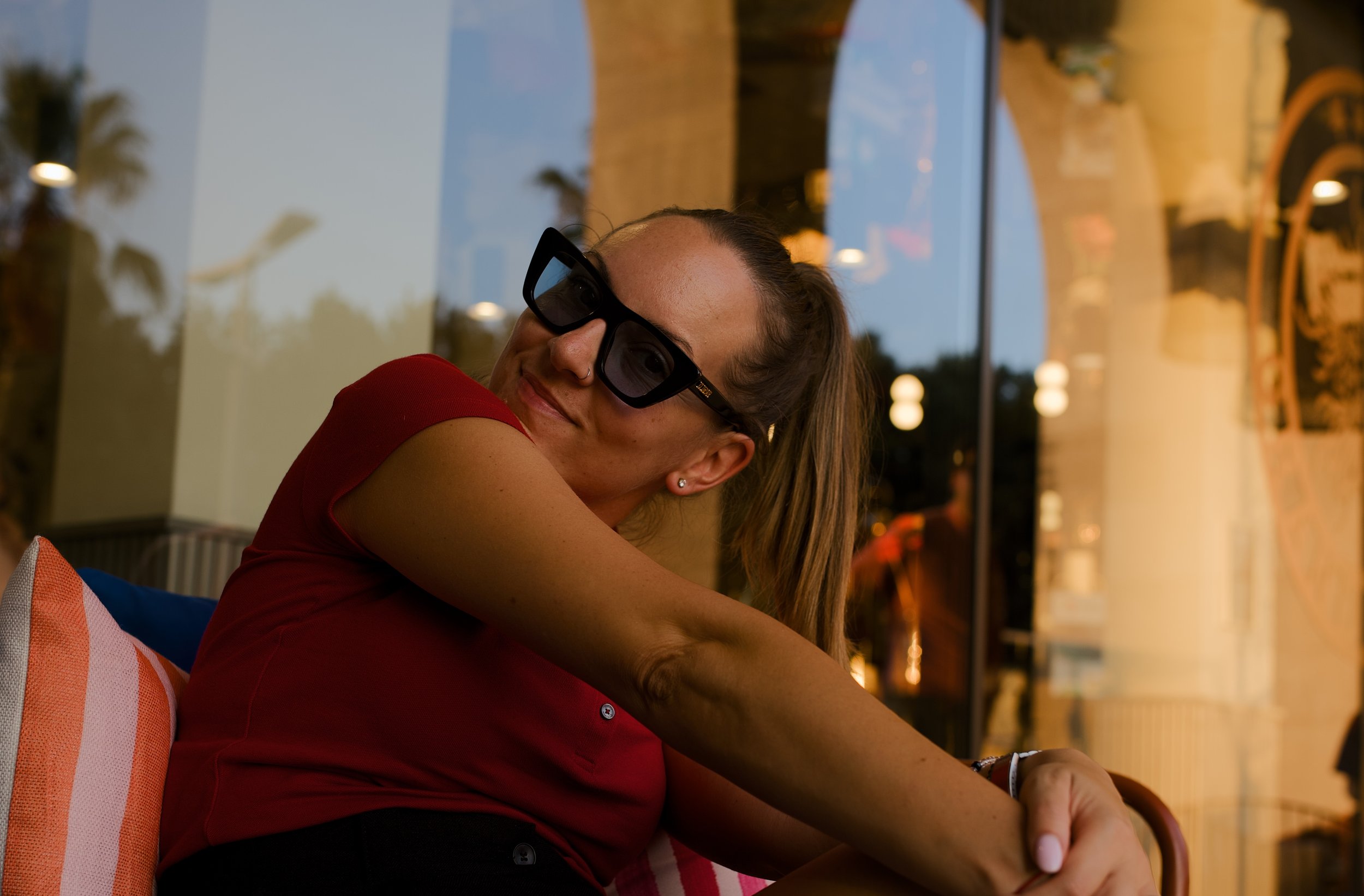 A woman with long hair sitting inside a restaurant, wearing sunglasses and a red shirt, with a view of the street and palm trees outside the window.