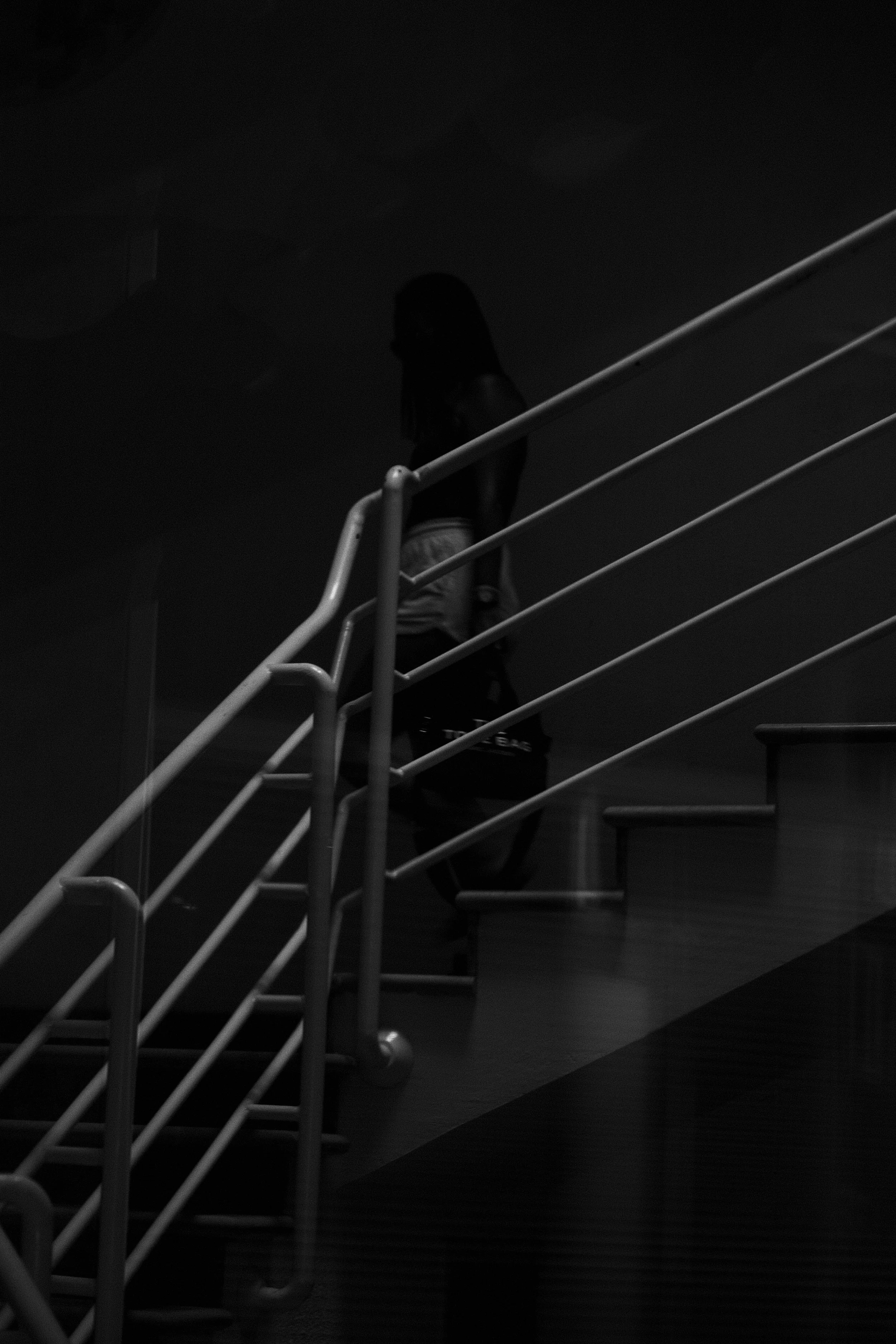 A person with long hair walking down a staircase with white railings, viewed from below, in a dark environment.