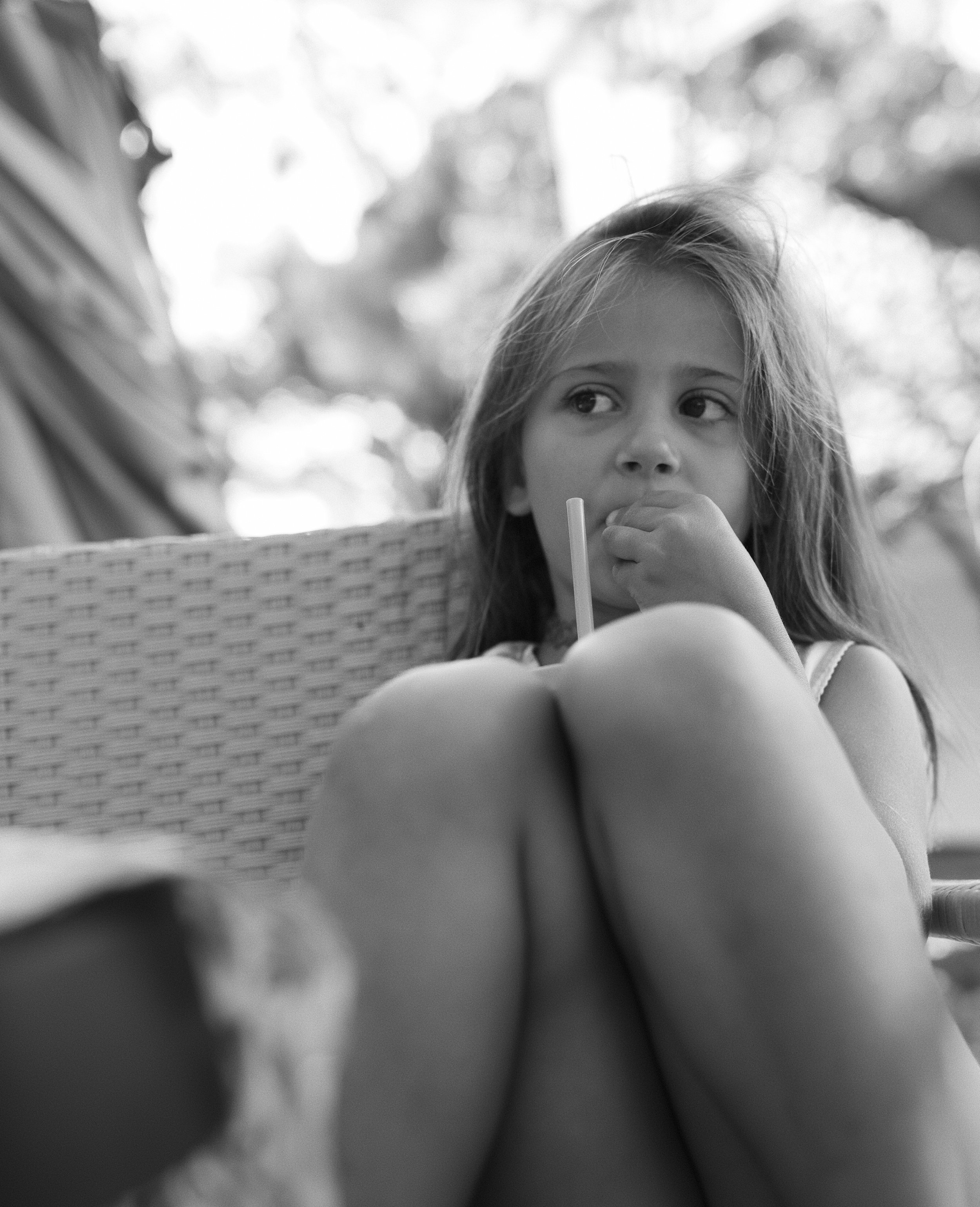 A young girl with long hair sitting outdoors, observed from a low angle in black and white, with blurred background and evident sunlight through trees.