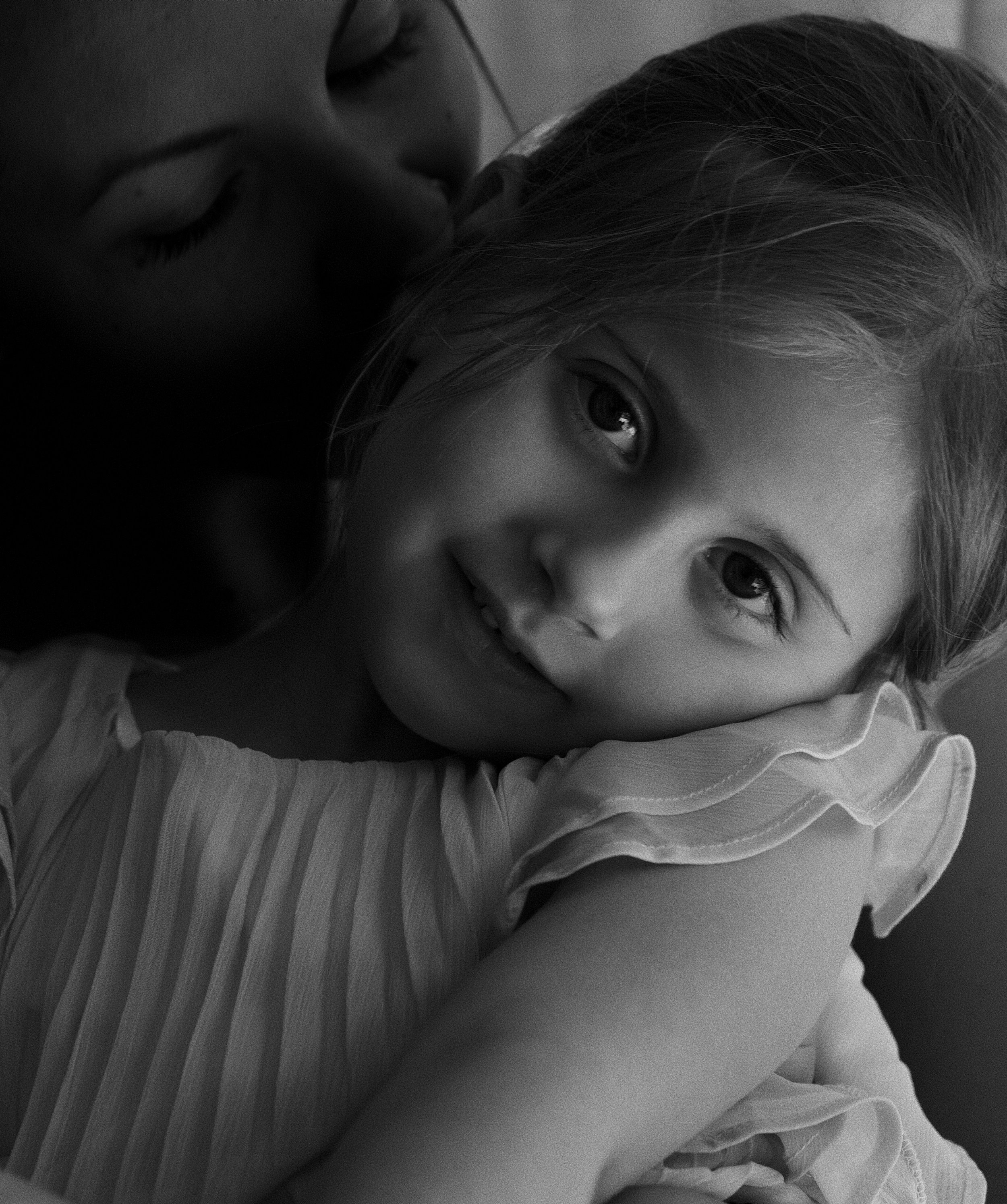 Close-up of a young girl with light-colored hair, large expressive eyes, resting her head on a pillow or surface, with a gentle expression, in black and white.